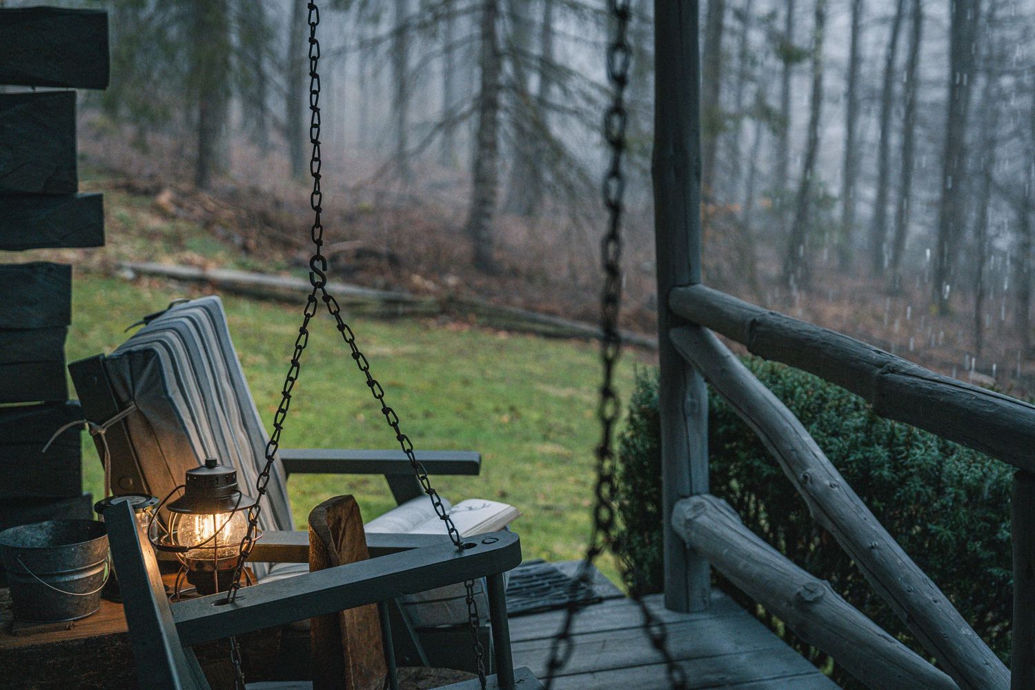 A cozy porch swing with glowing lantern and book sits on a misty evening deck overlooking a foggy forest.