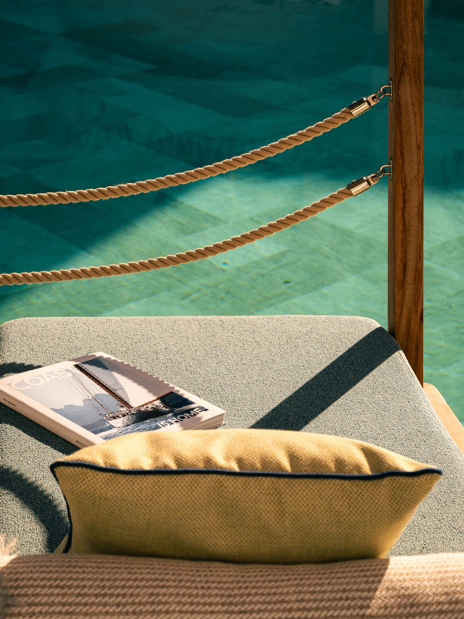 Beige cushions and book arranged on deck beside turquoise water with rope railing.