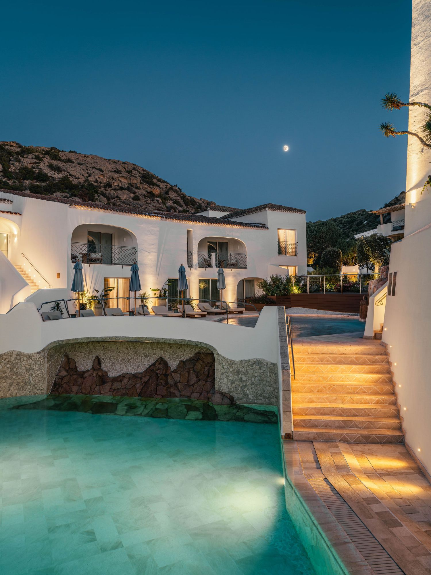 Illuminated resort swimming pool with white buildings and stone steps at twilight.