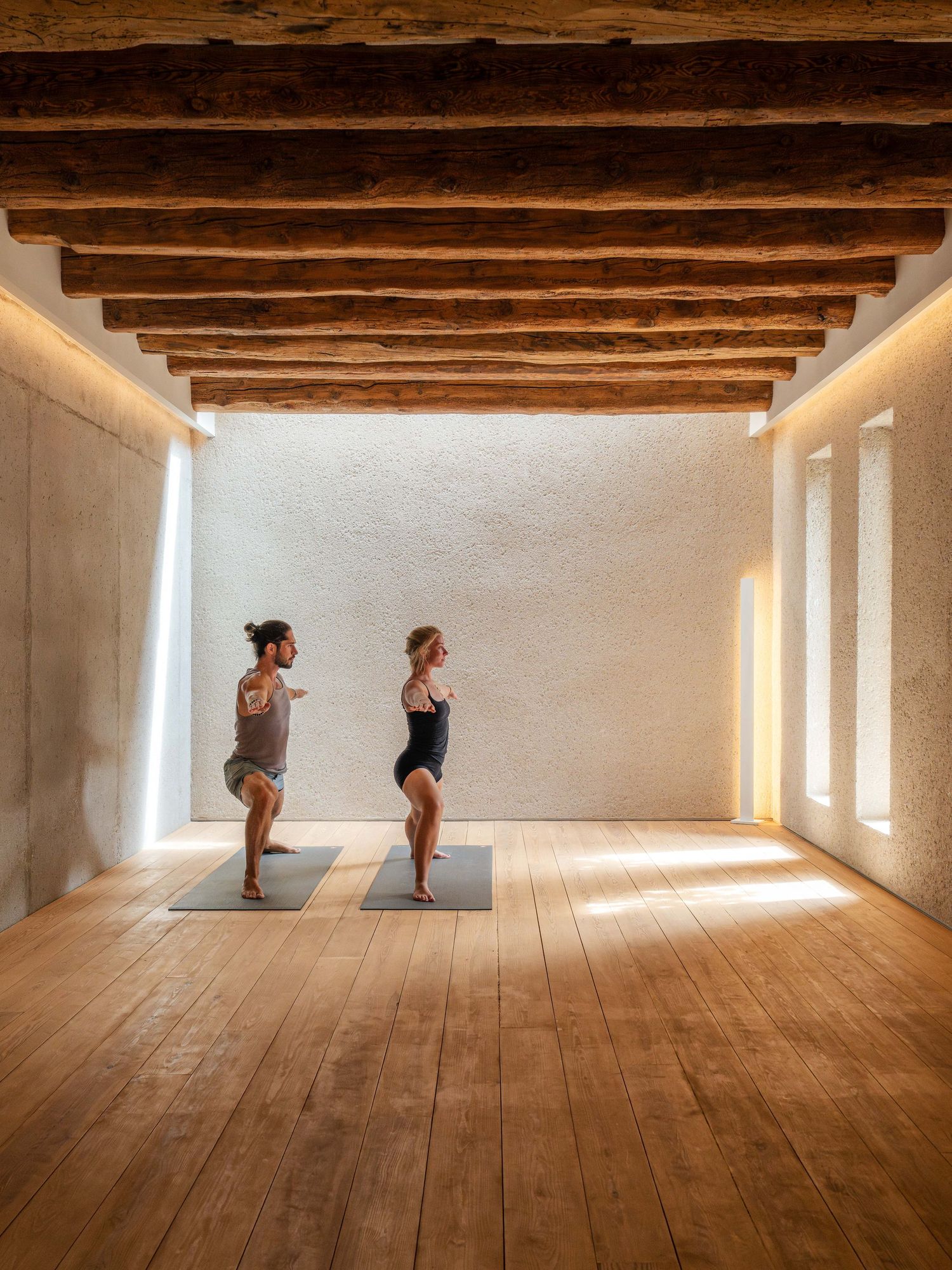 Two people practicing yoga handstands against a white wall in a rustic room with wooden beams.