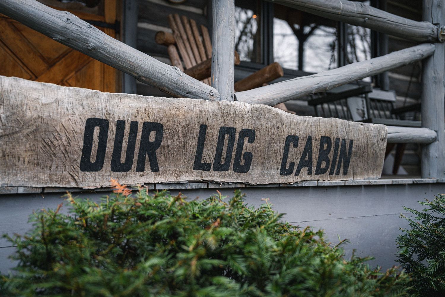 A rustic wooden sign for 'Our Log Cabin' appears weathered against a grey metal background and natural outdoor setting.