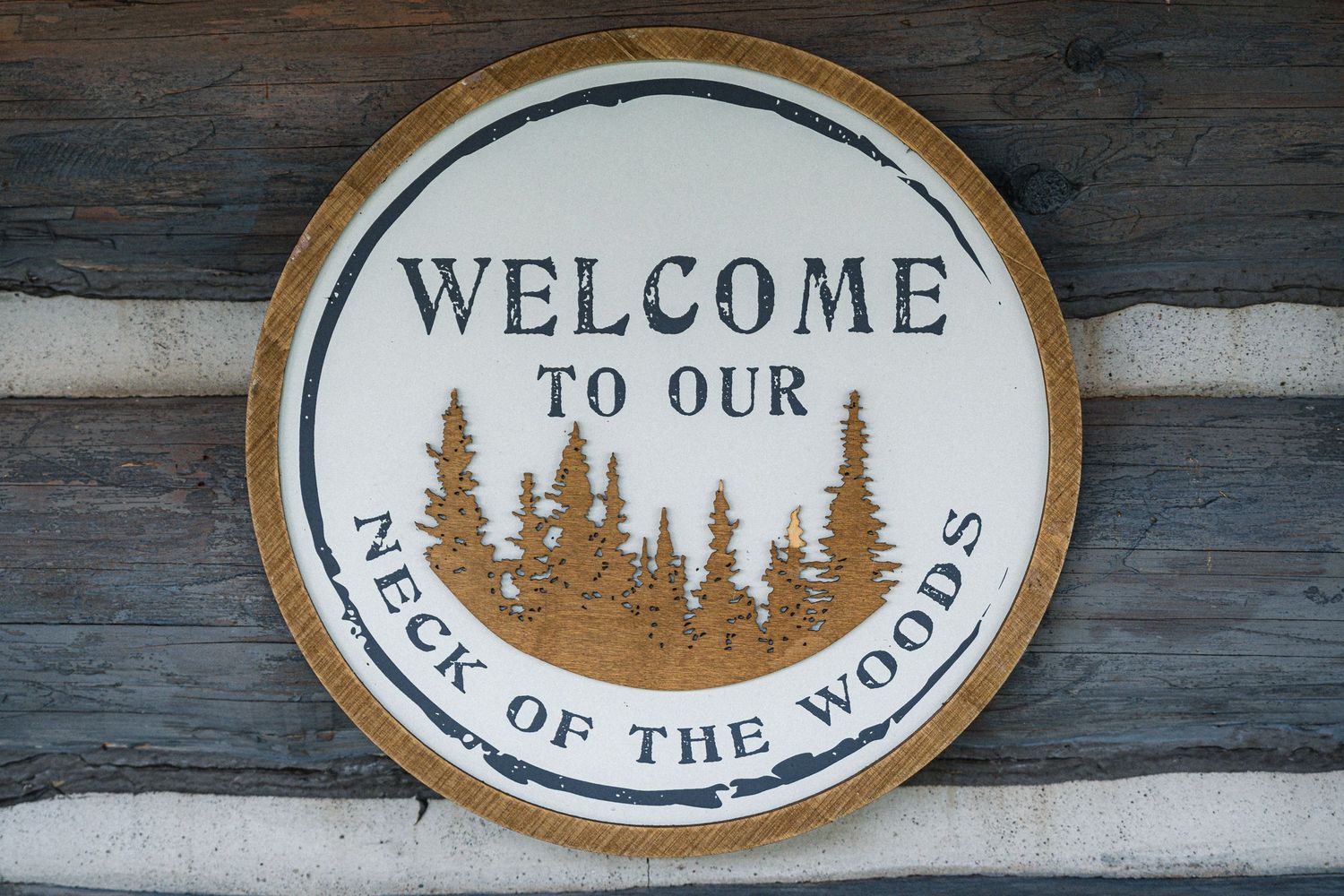 A round wooden welcome sign with pine tree silhouettes reading 'Welcome to our neck of the woods' mounted on log cabin wall.
