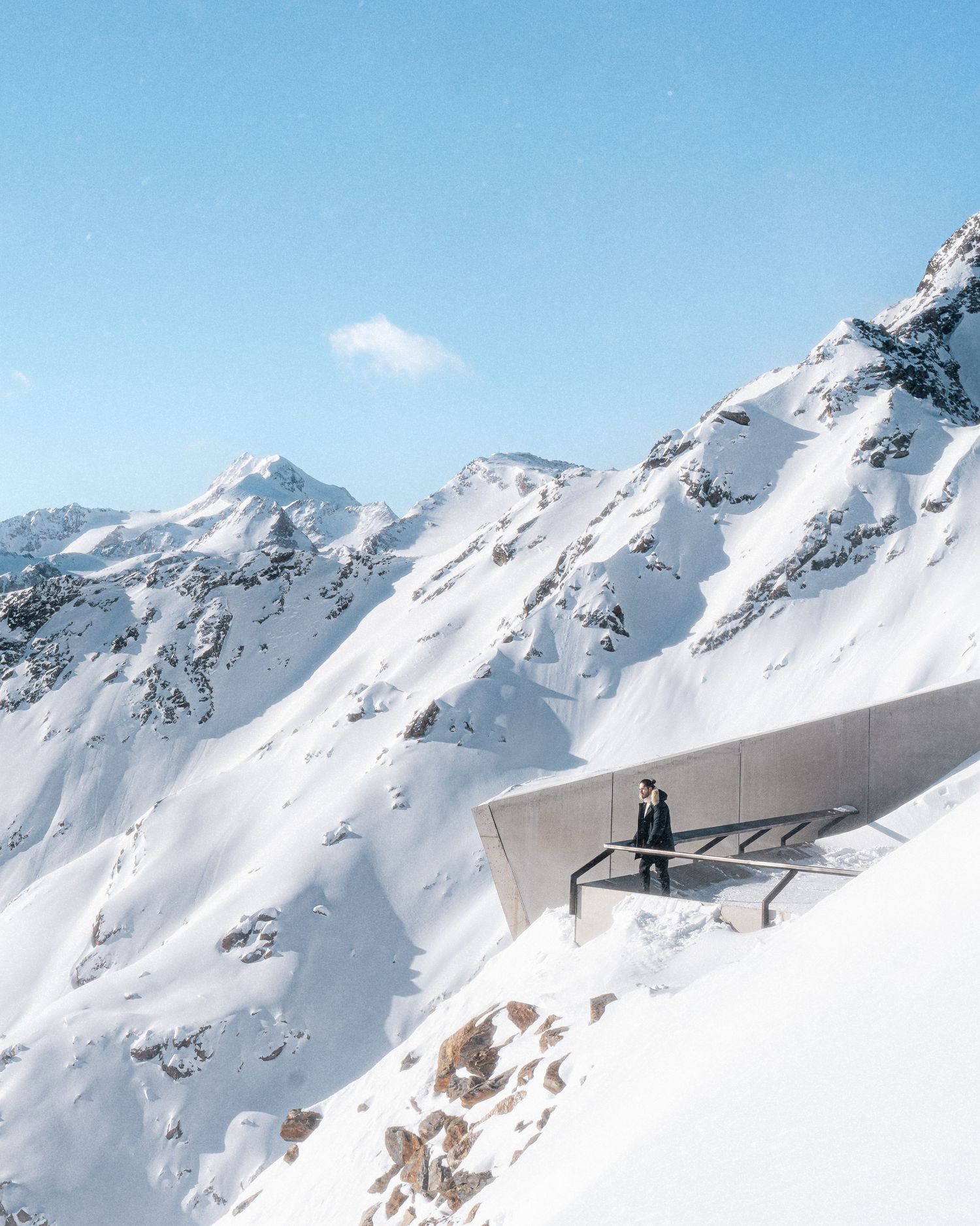 Snow-covered mountain peaks with a modern glass viewing platform extending over the dramatic winter landscape.