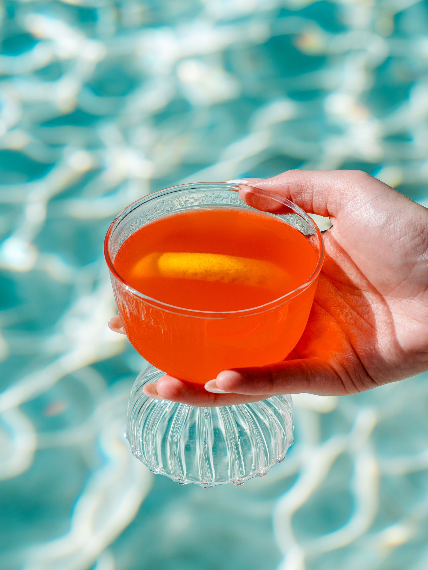 Orange tropical cocktail in a clear glass held against sparkling pool water background.