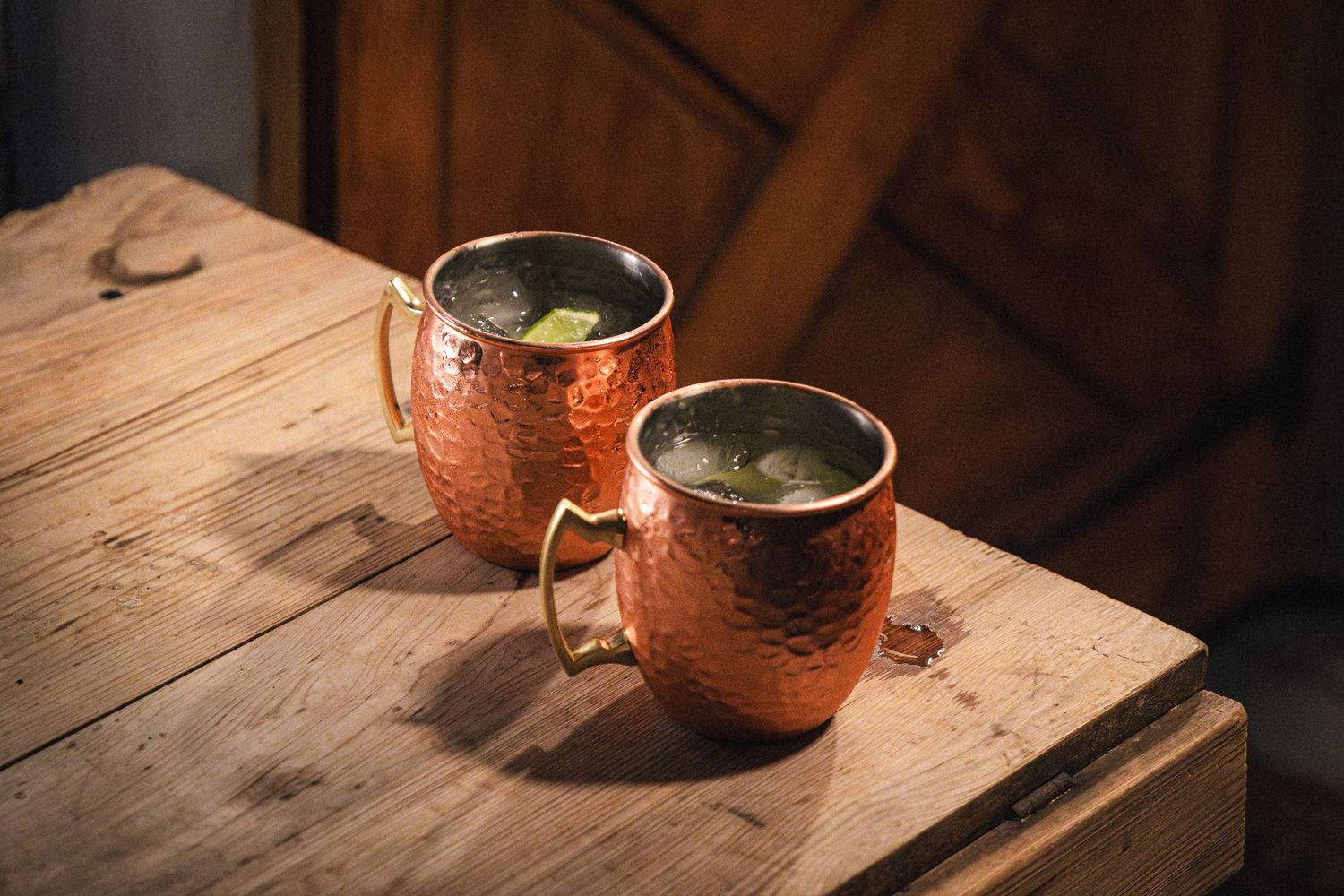 Copper Moscow mule mug with lime garnish sits on rustic wooden surface in warm lighting.