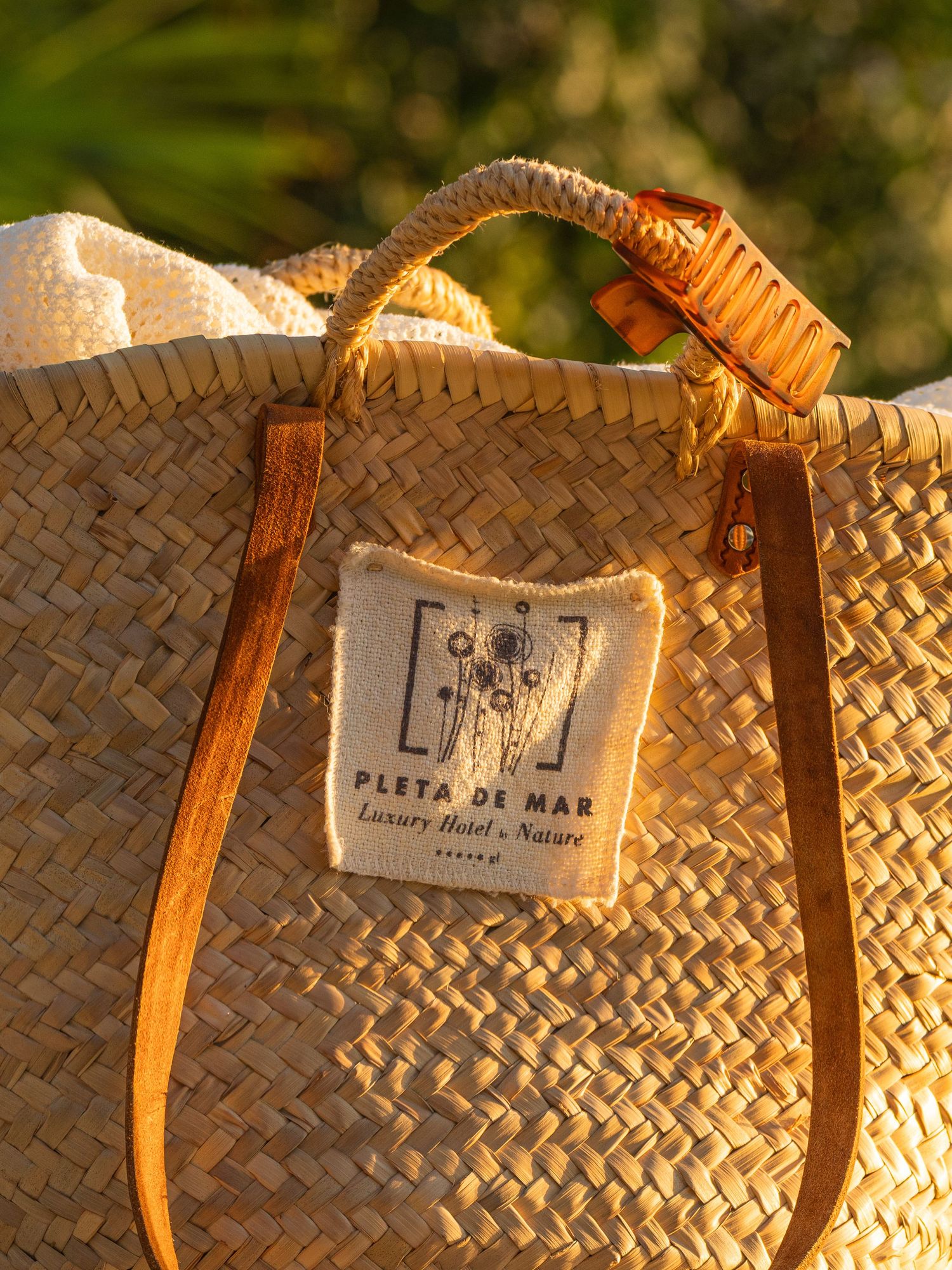 Close-up of a woven straw beach bag with leather handles in golden sunlight.