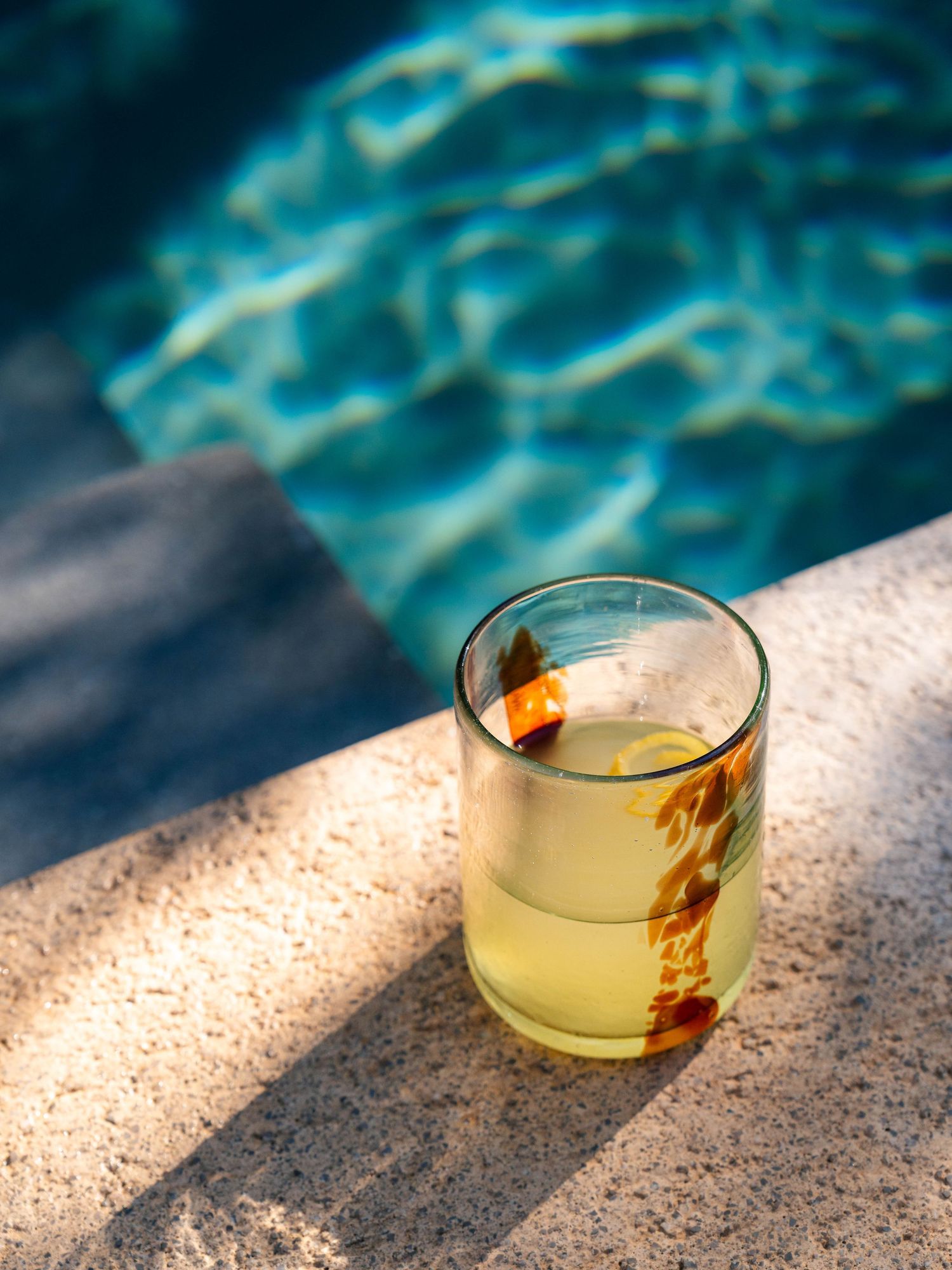 Glass of clear cocktail with garnish sitting by turquoise pool water in sunlight.