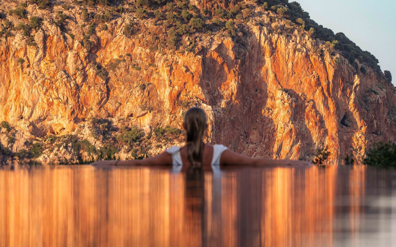 Dramatic sunset reflection on rocky cliffs along a calm waterfront creating golden orange hues.