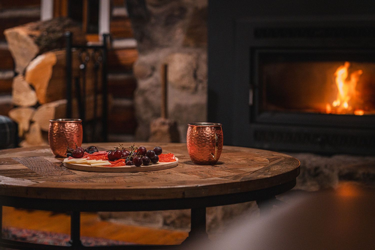 A cozy fireplace scene with two copper mugs and snacks on a rustic wooden table.