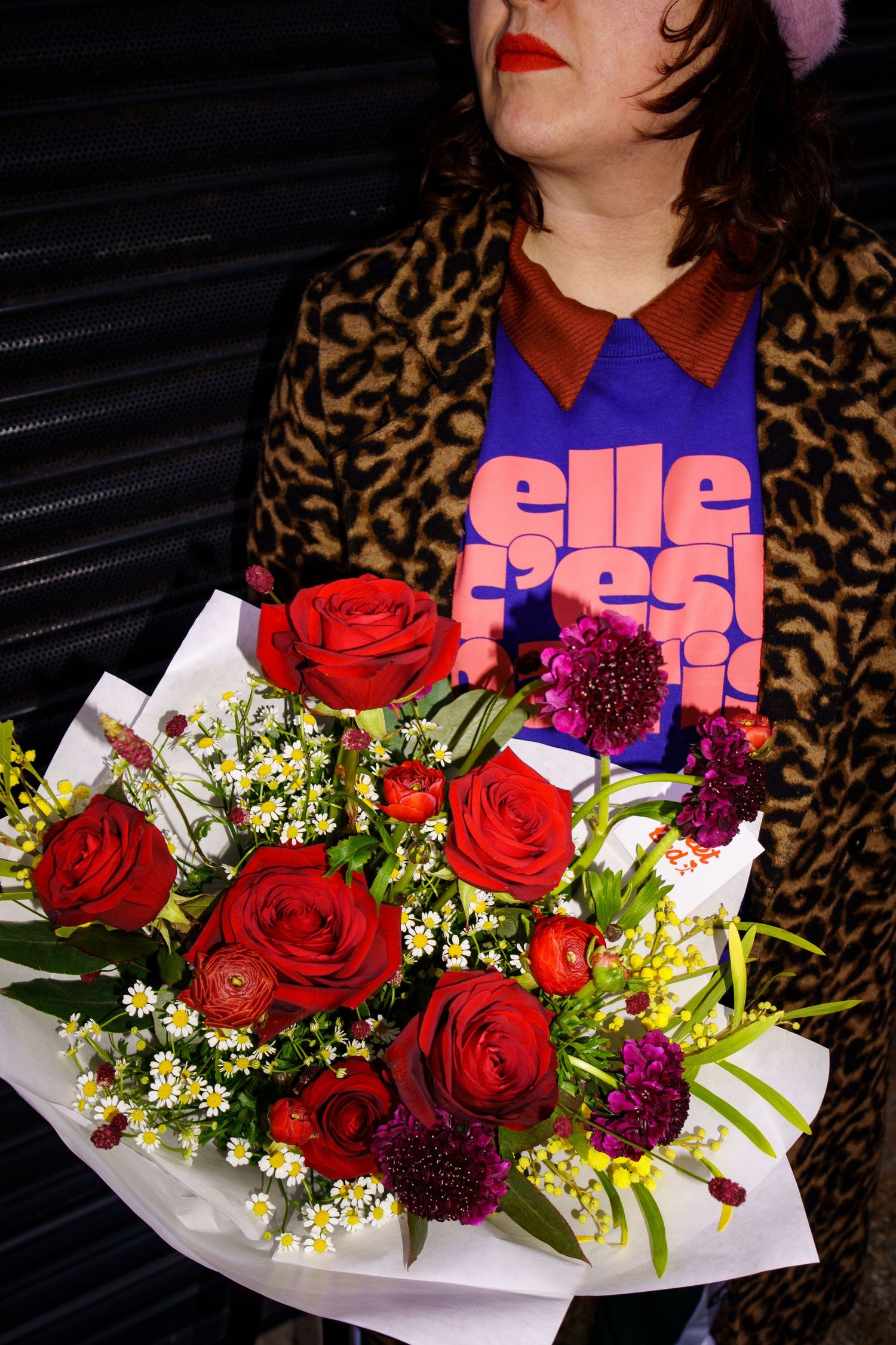 A vibrant bouquet of red roses and purple flowers held against a leopard print coat and colorful graphic t-shirt.