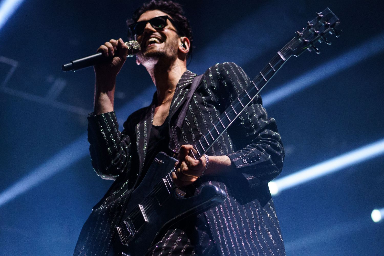 Rock musician performing on stage, playing electric guitar and singing into microphone, wearing pinstripe suit under blue stage lights.