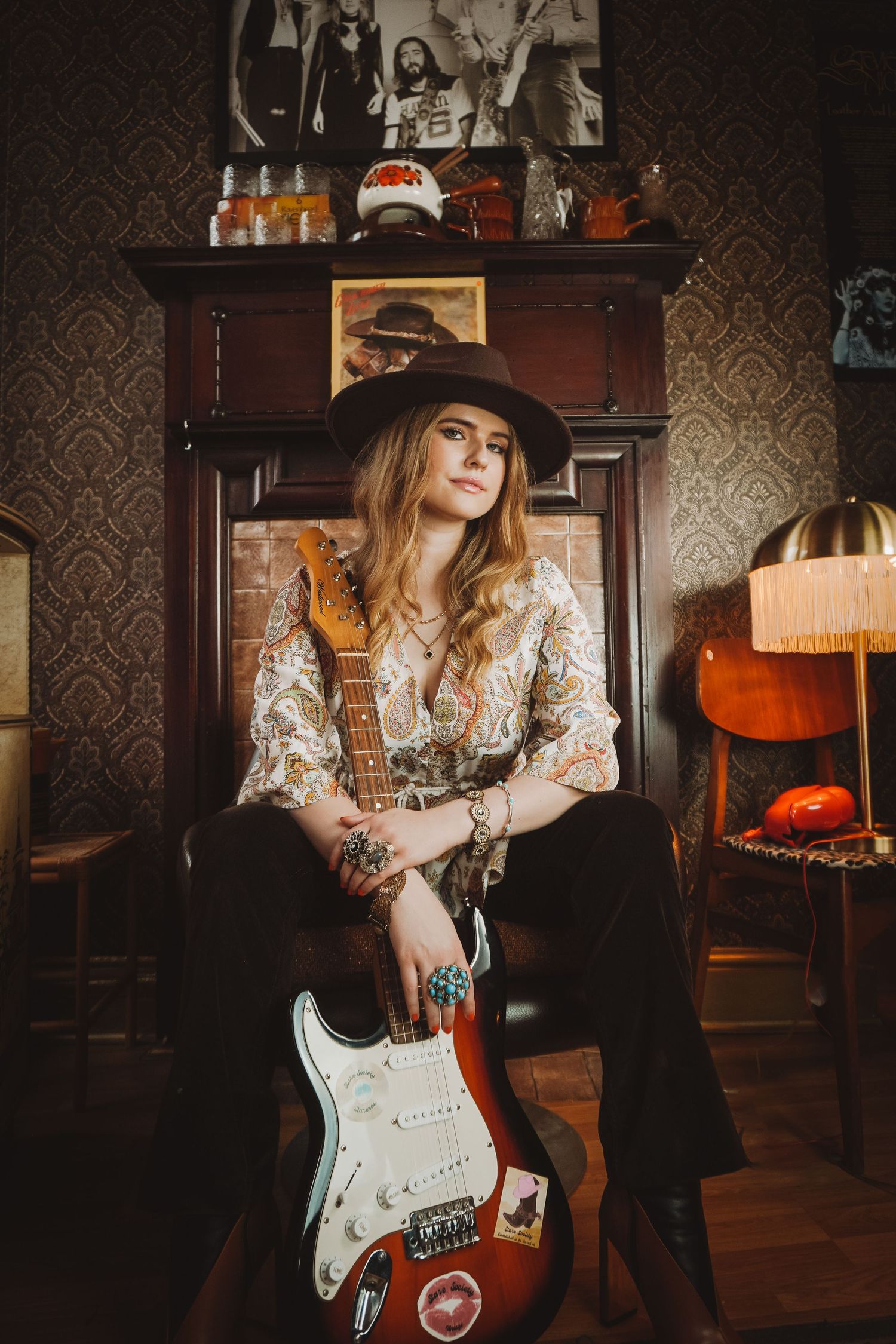 Musician in vintage-style lace playing electric guitar in a dark room with patterned wallpaper.