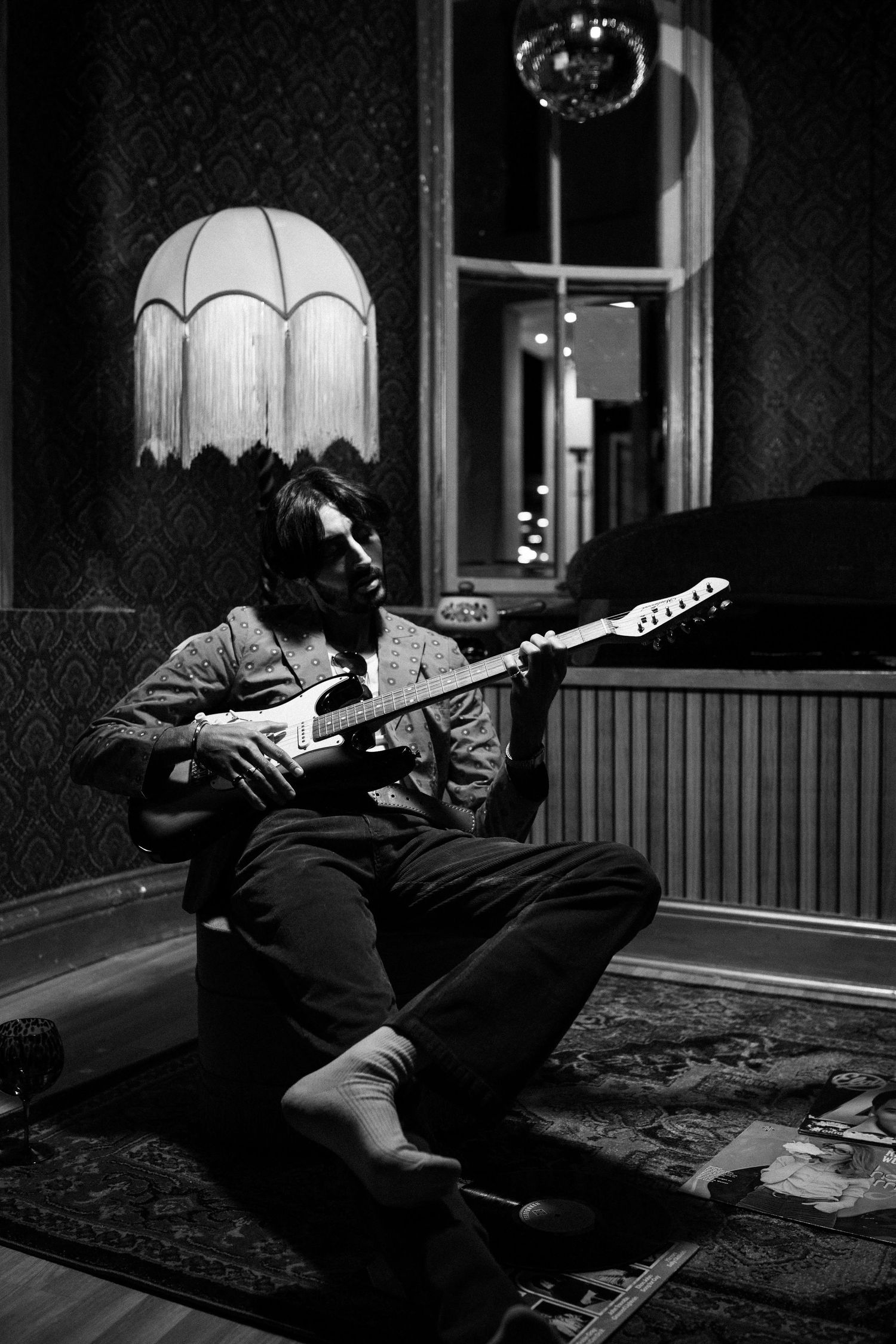 Black and white photo of guitarist playing on floor beneath vintage lampshade.