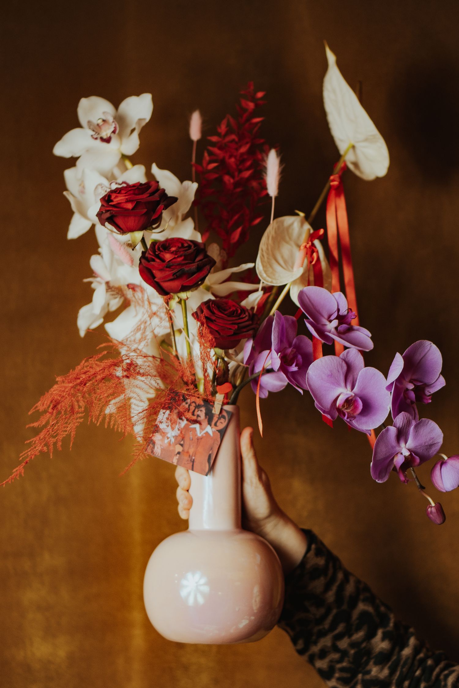 An artistic still life of red roses and purple orchids in a pink vase against a warm brown background.