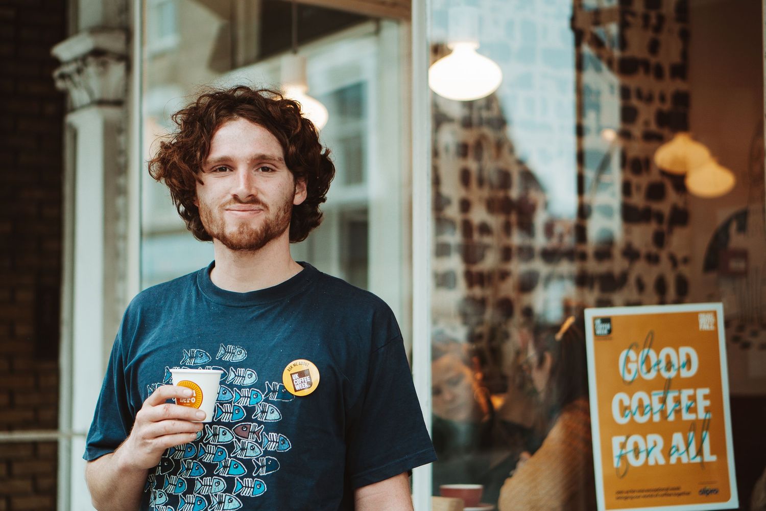 A person in a dark t-shirt holds a coffee cup while standing outside a cafe with a 'Good Coffee For All' sign.