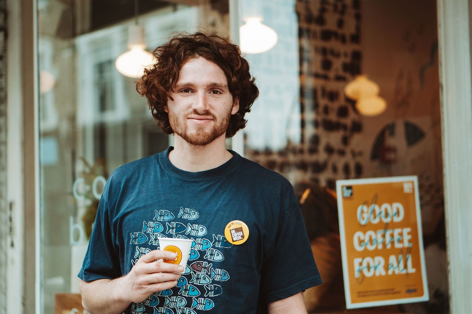 A casual portrait of someone smiling outside a coffee shop while holding a beverage.