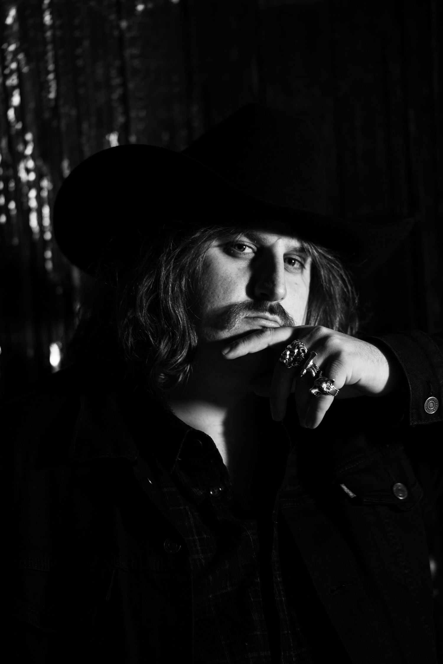 A moody black and white portrait of a person wearing a dark hat and rings in low-key lighting.