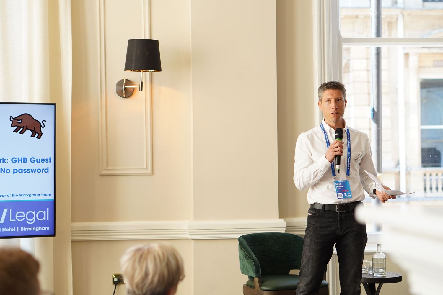 Speaker presenting to an audience at a business conference in a well-lit room with cream-colored walls.
