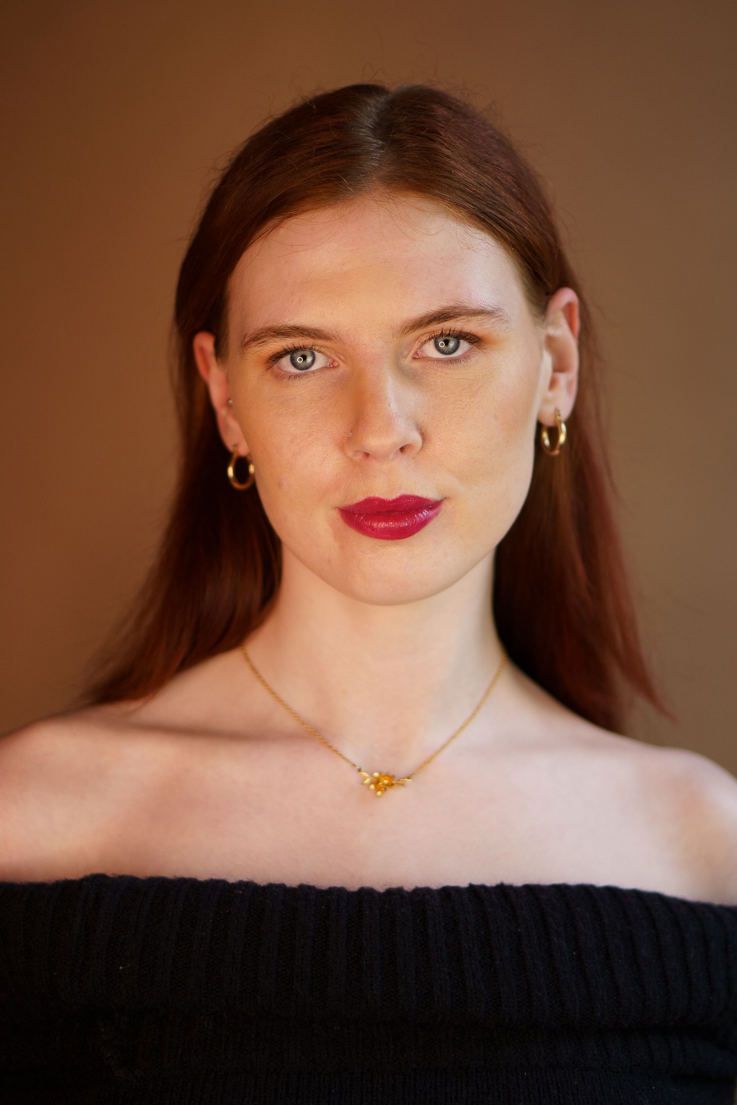 Portrait against brown background wearing black off-shoulder top, red lipstick and delicate gold necklace.