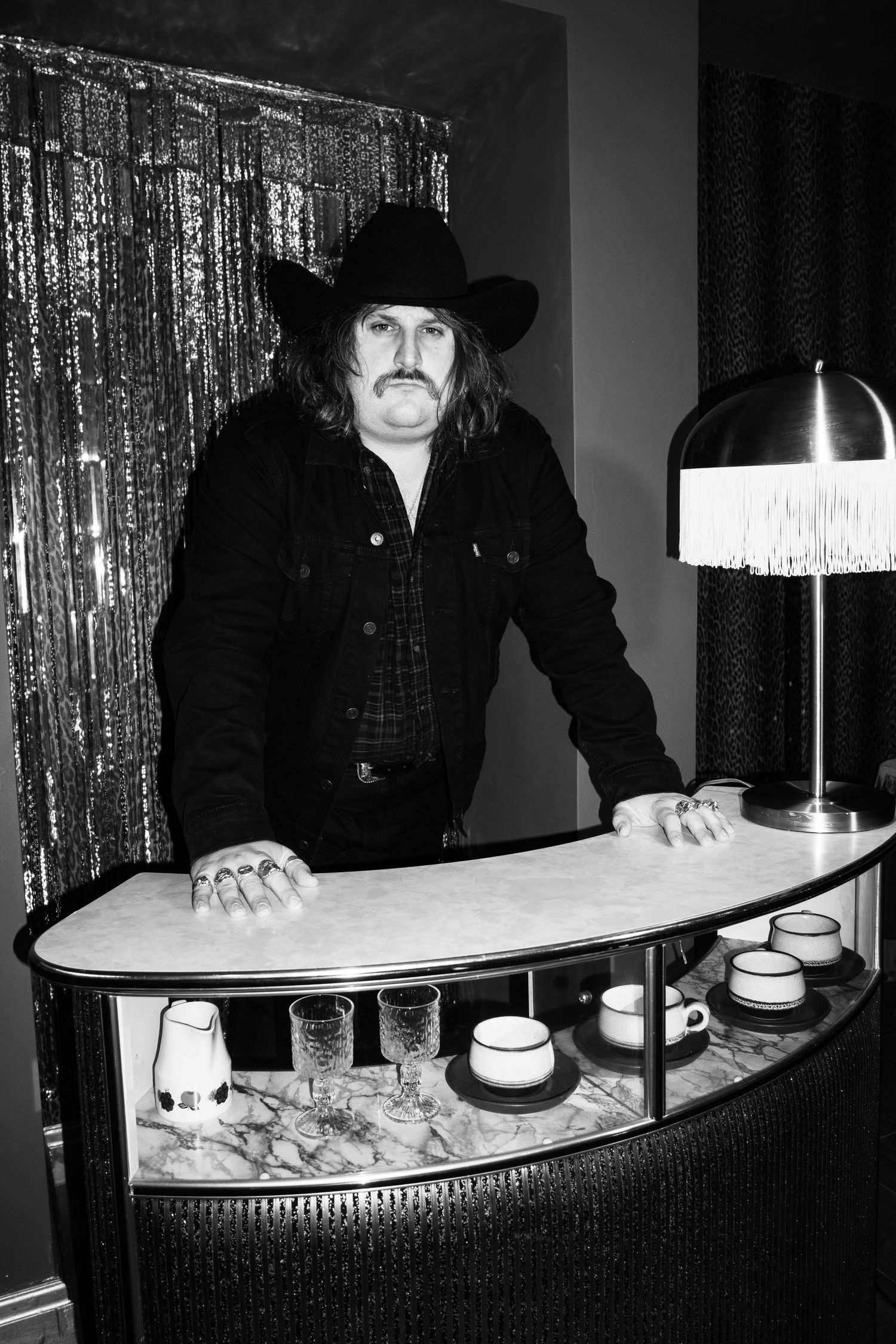 Black and white photo of a man in cowboy hat leaning on a vintage bar display case with lamp and glassware.