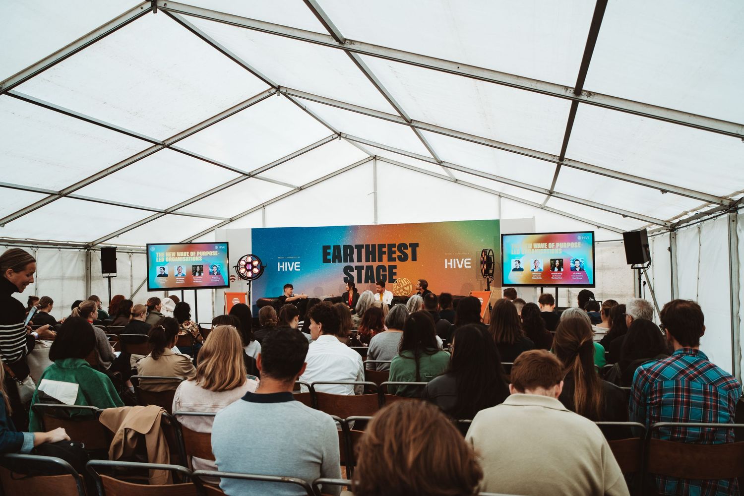 A crowded audience sits in rows inside a white event tent with branded banners visible in the background.