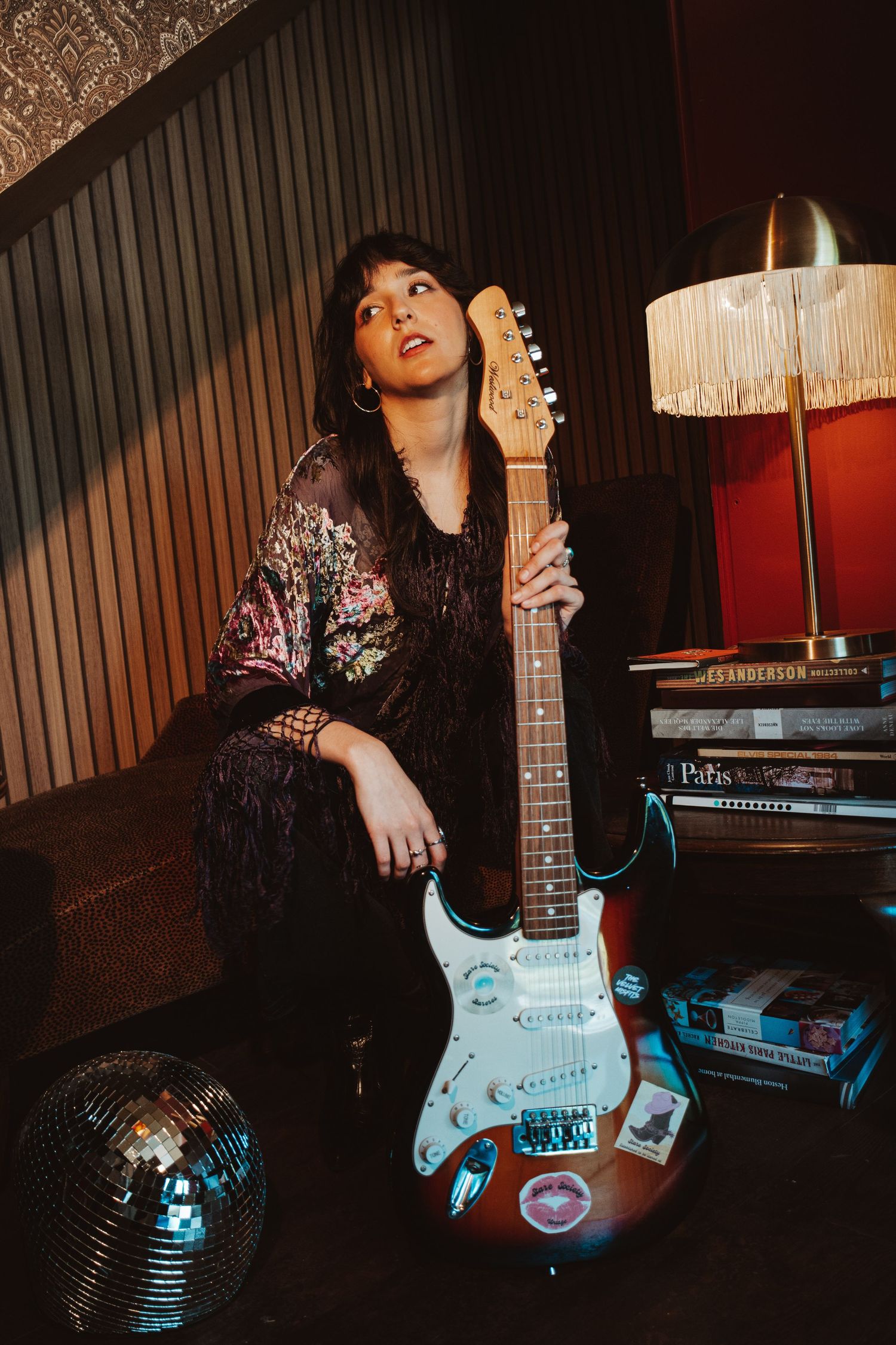 A moody portrait of a musician with a Fender Stratocaster guitar in a vintage-styled room with disco ball.