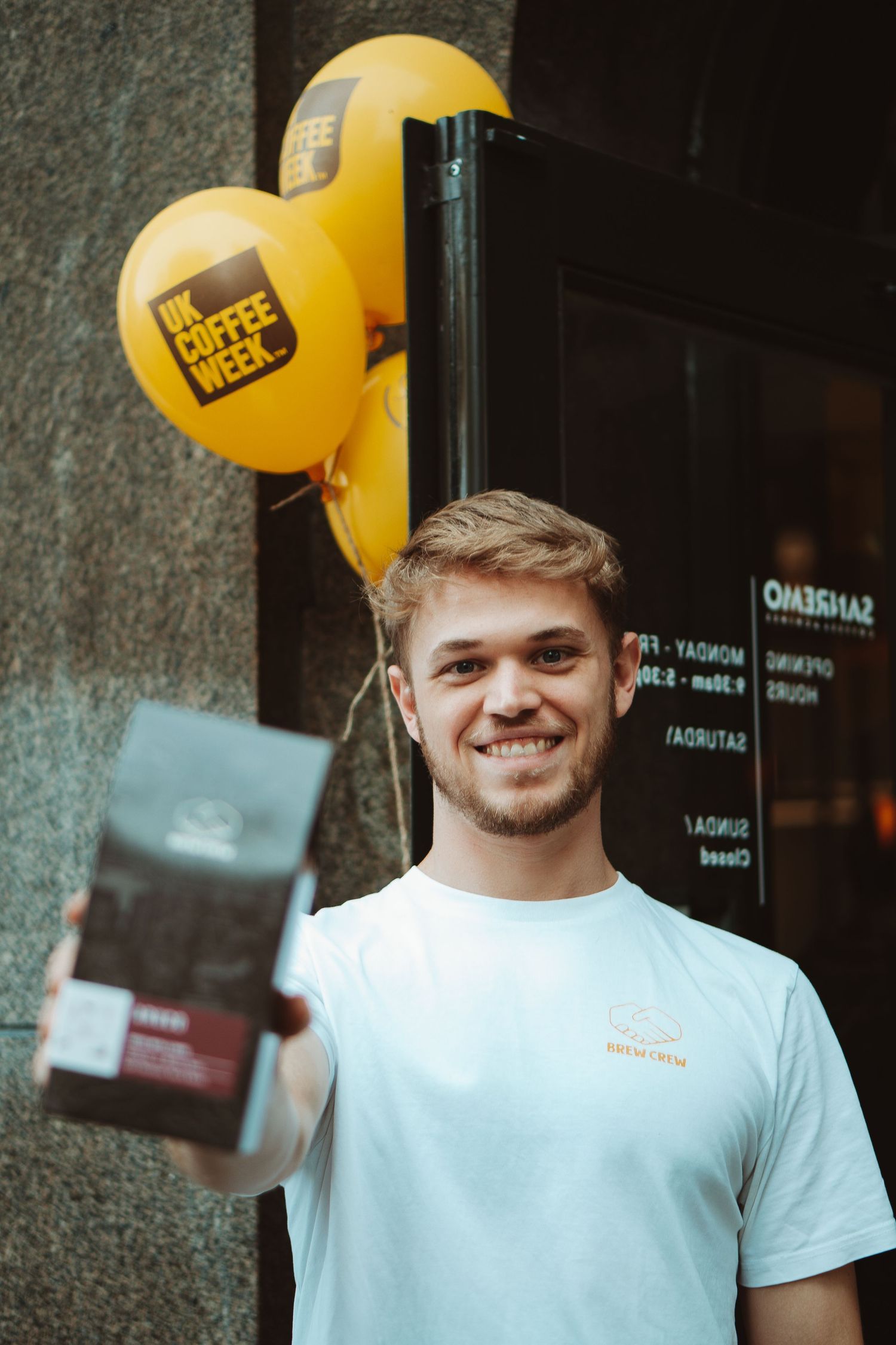 Person in white t-shirt smiling with yellow balloon against dark wall background.