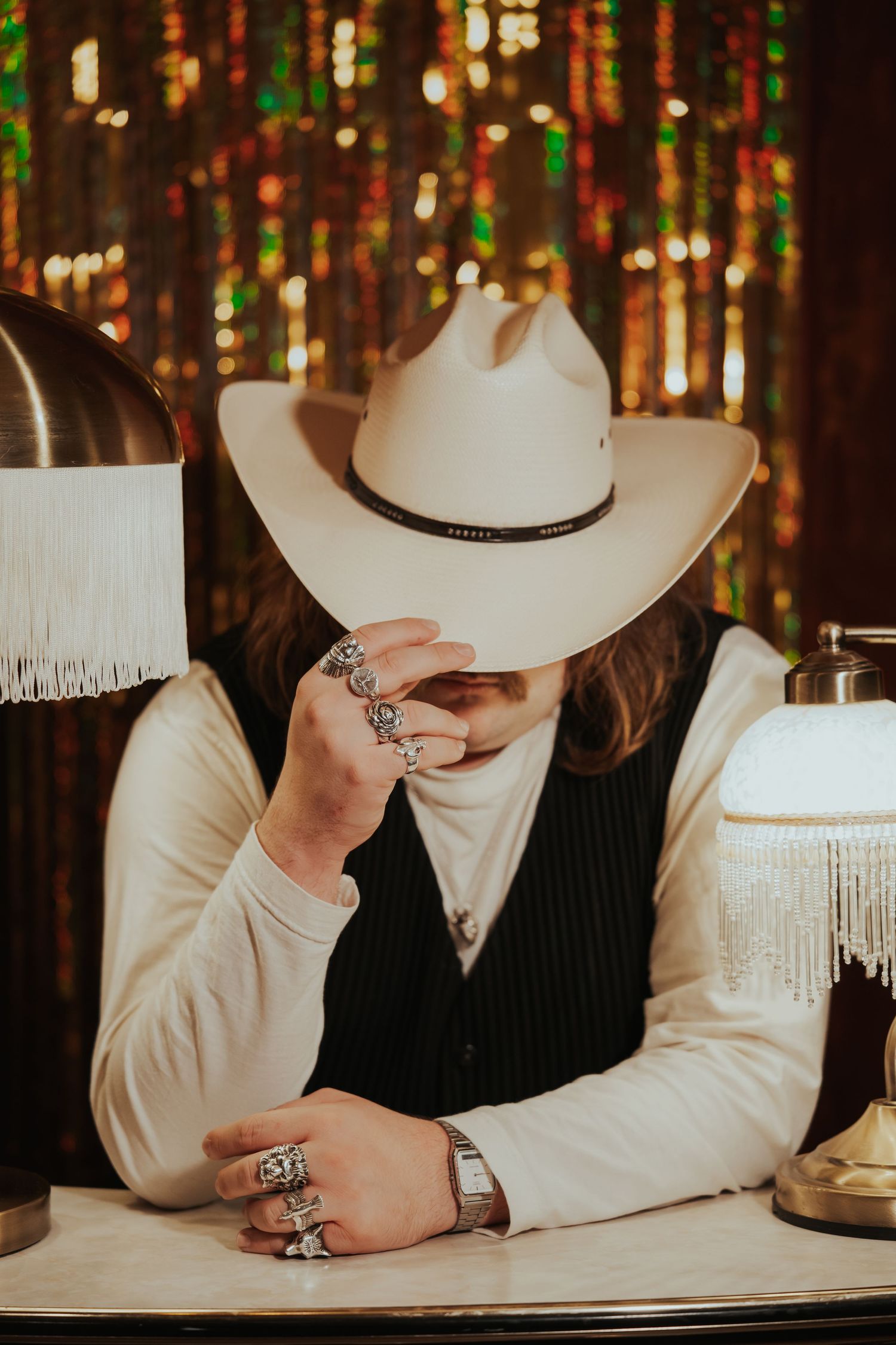 Person in white cowboy hat and rings sits at desk before glittering backdrop with fringed lamps.