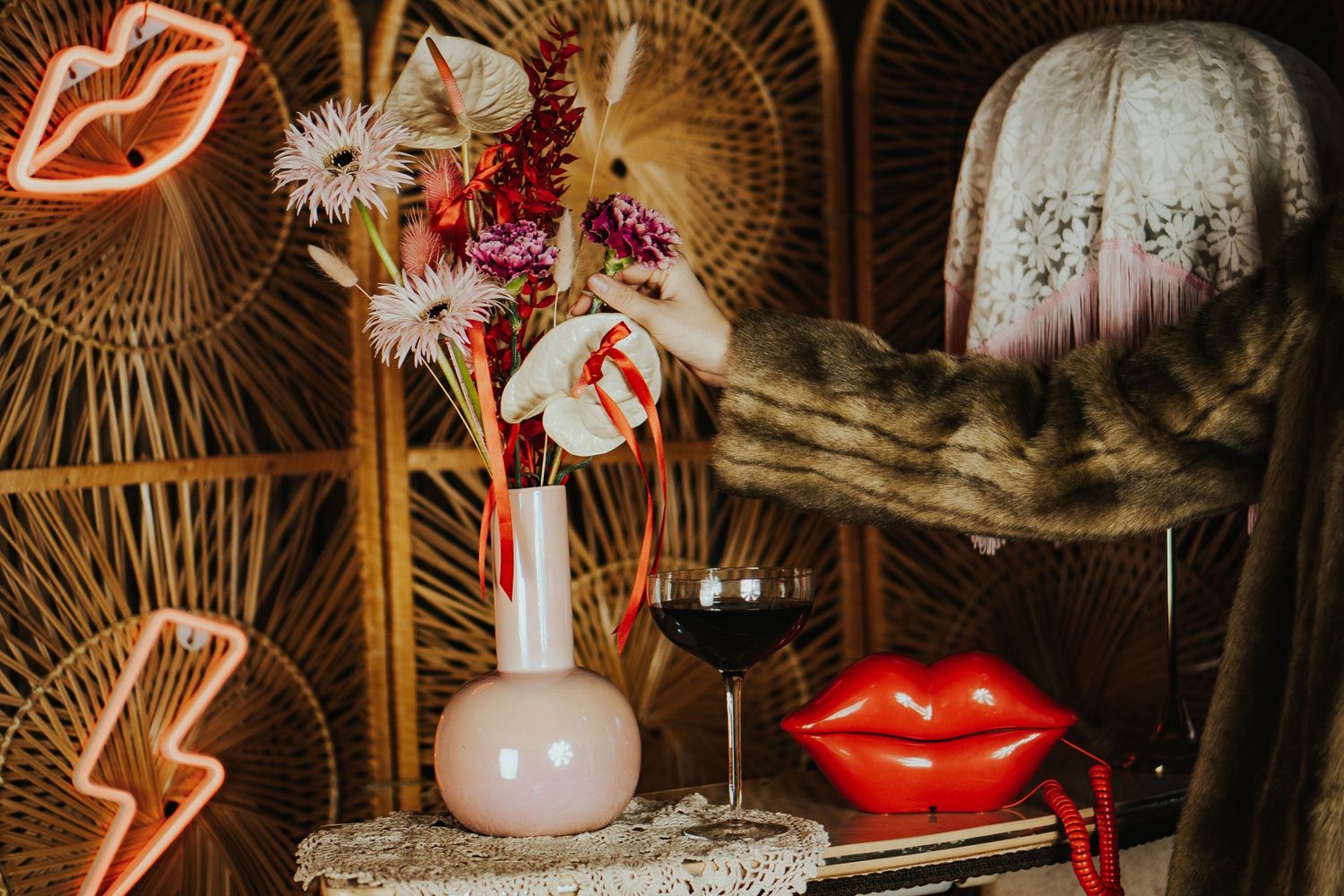 Retro styled still life with neon lips sign, wine glass, white vase with flowers, and decorative pumpkin against rattan backdrop.