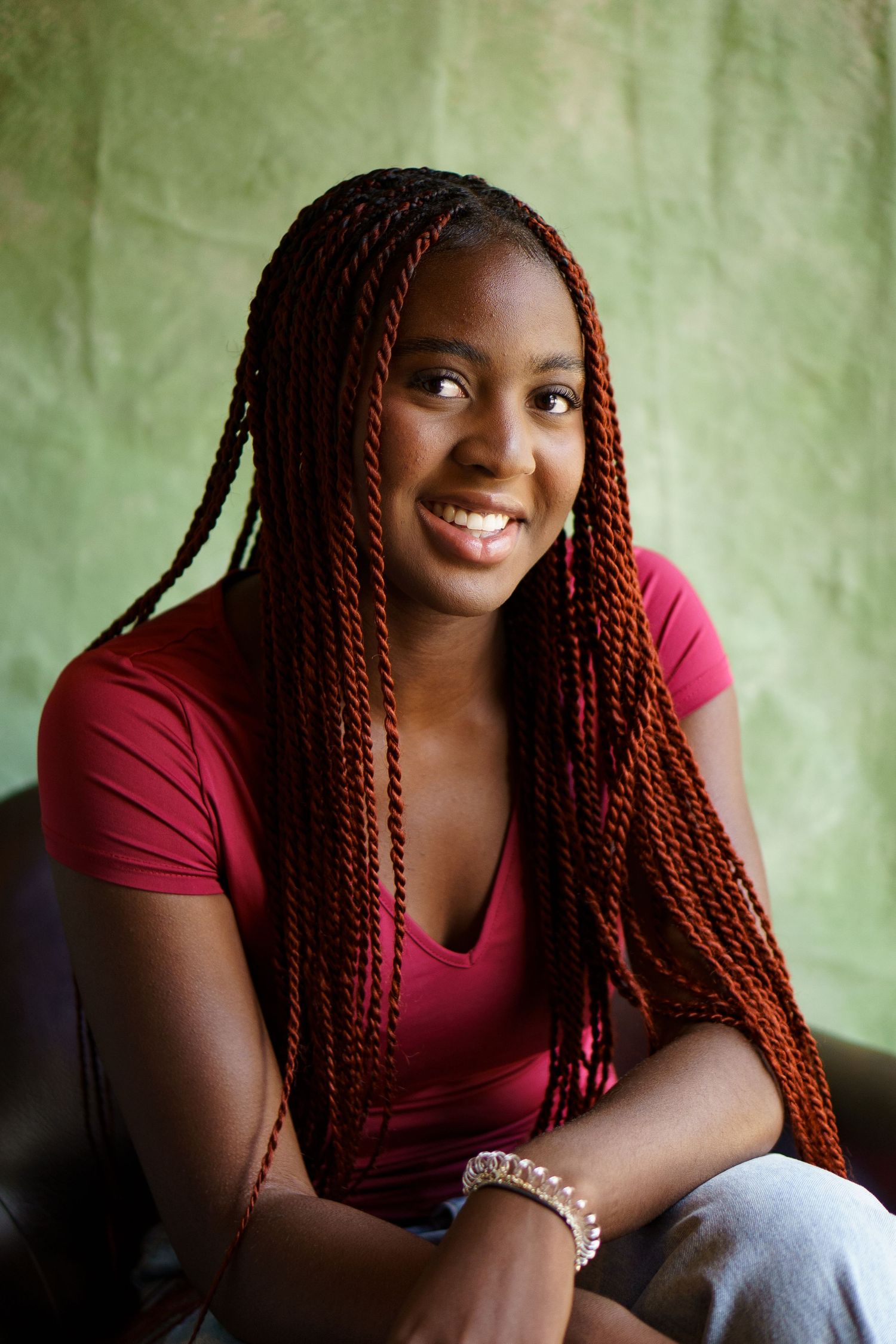 A person with red braids wearing a burgundy top sits casually against a green textured wall.
