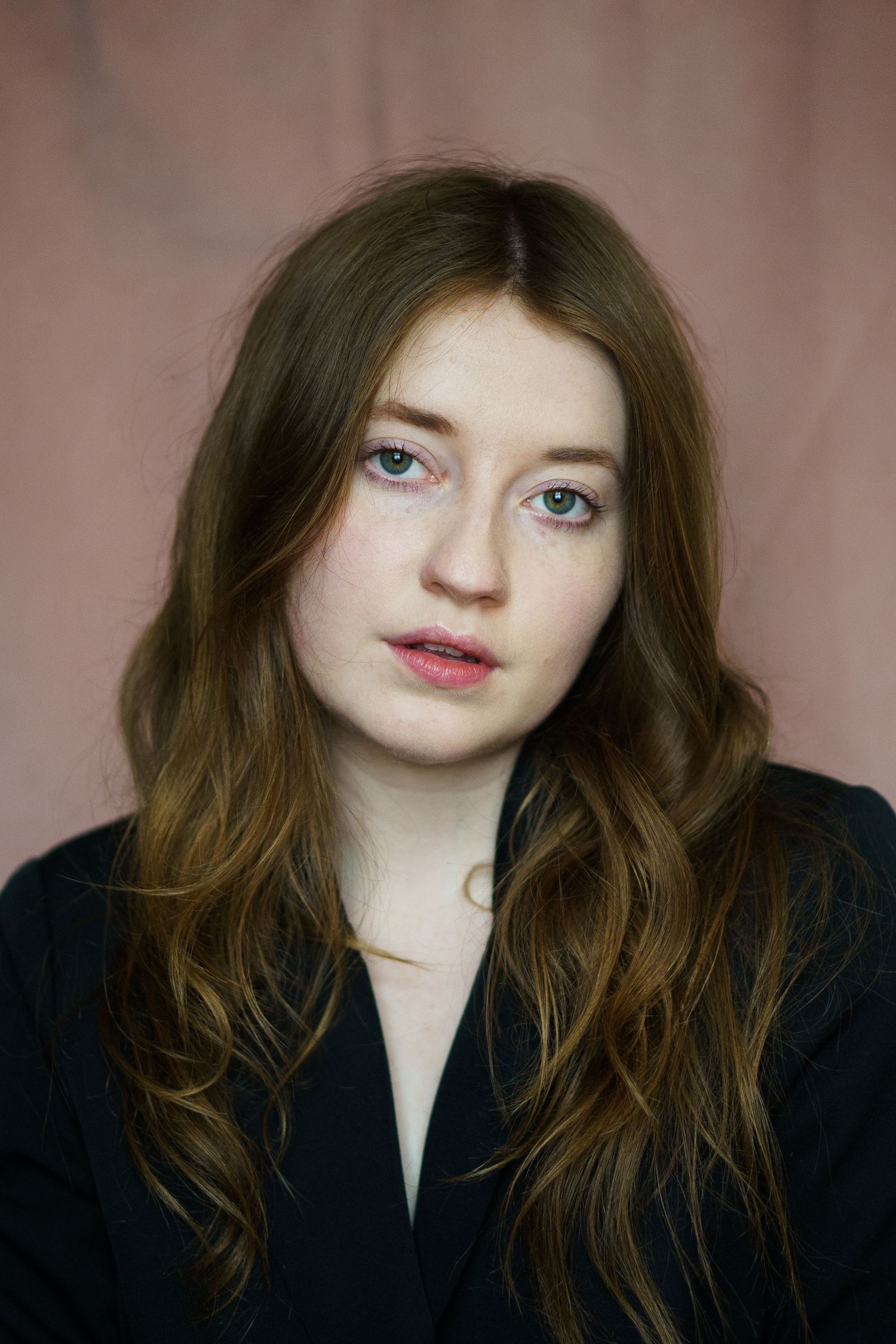 Young woman with long auburn hair, freckles, and blue eyes wearing a black jacket against a pink background.