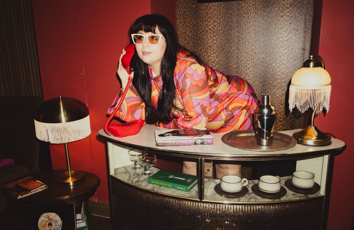 Retro lounge scene with vintage lamps and coral-colored dress against patterned wallpaper.