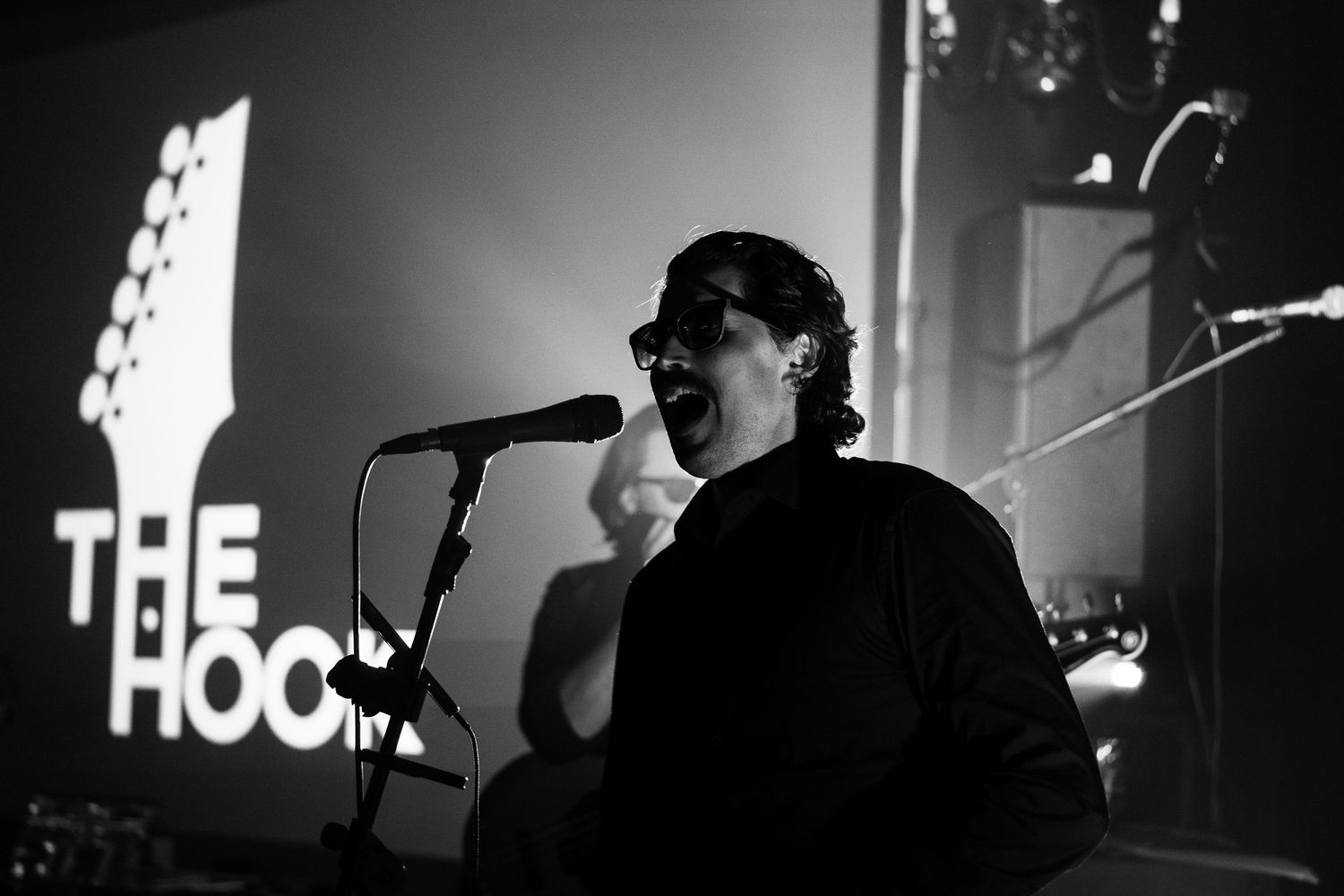 Black and white photo of a singer performing at The Hook venue, speaking into a microphone on a dimly lit stage.