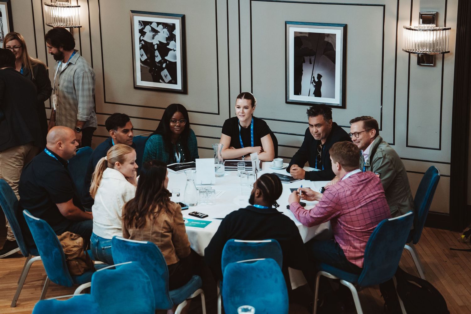 A group of people sit around a round table in a meeting room with blue chairs and framed artwork on the walls.