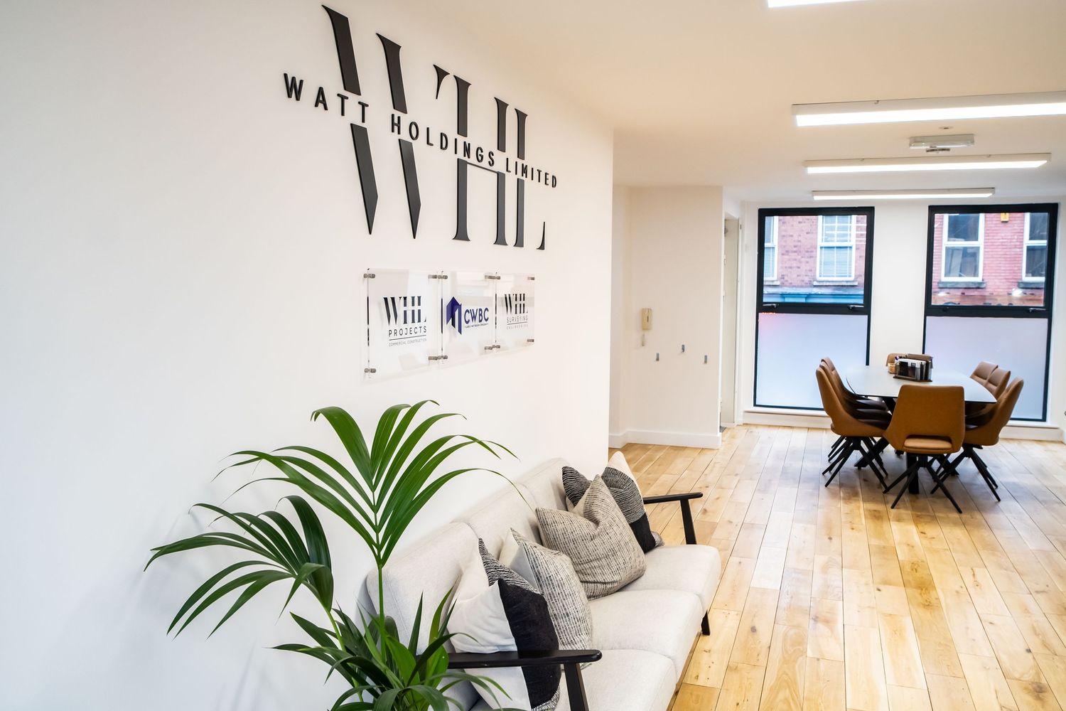 Modern open office space with hardwood floors, white walls, and potted plants features natural lighting and minimalist decor.