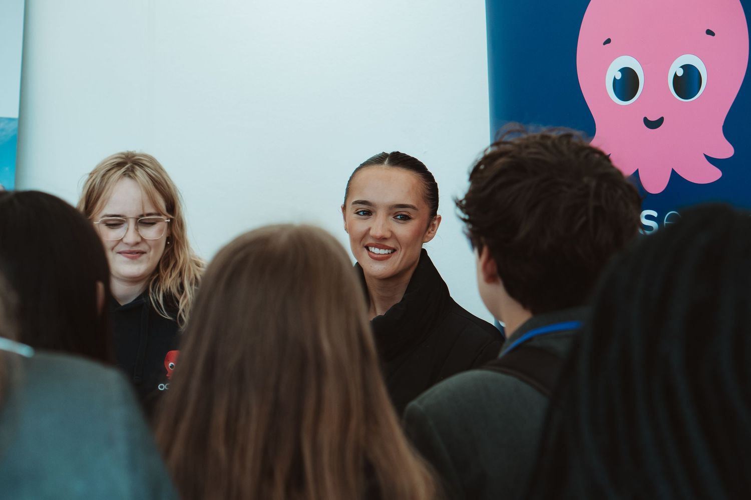 A group of people gather in a room with a pink cartoon octopus decoration visible on the wall.