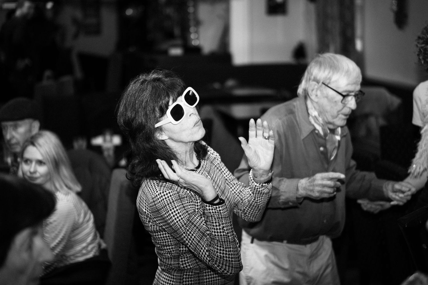 A black and white photo of people dancing, with one wearing white sunglasses and a checkered jacket.