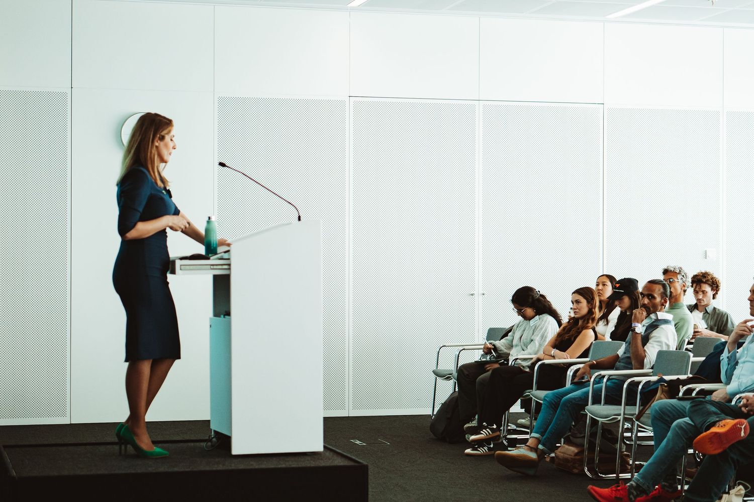 A speaker in business attire presents from a podium to an audience seated in rows during a professional conference.