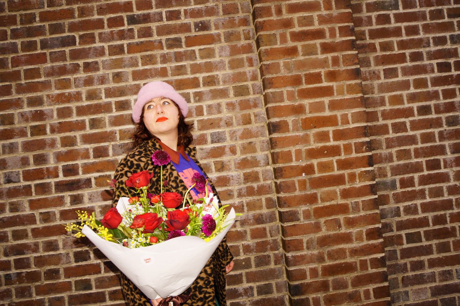 A person in winter attire holds a vibrant bouquet of red and yellow flowers against a brick wall background.