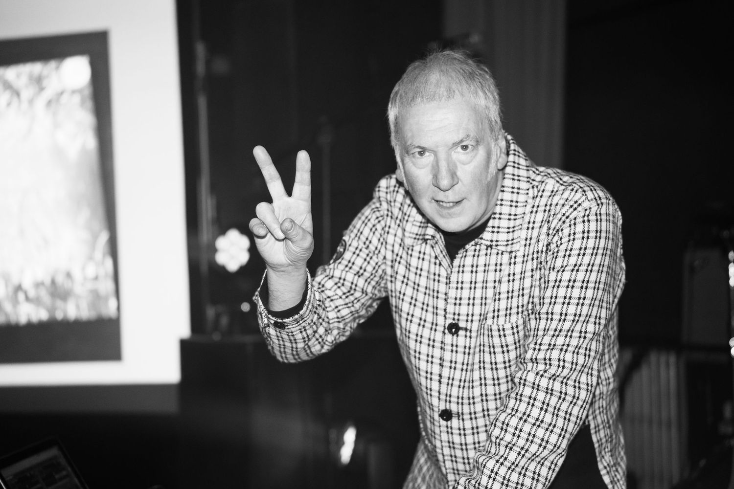 Black and white photo of a smiling man in a plaid blazer making a peace sign gesture indoors.