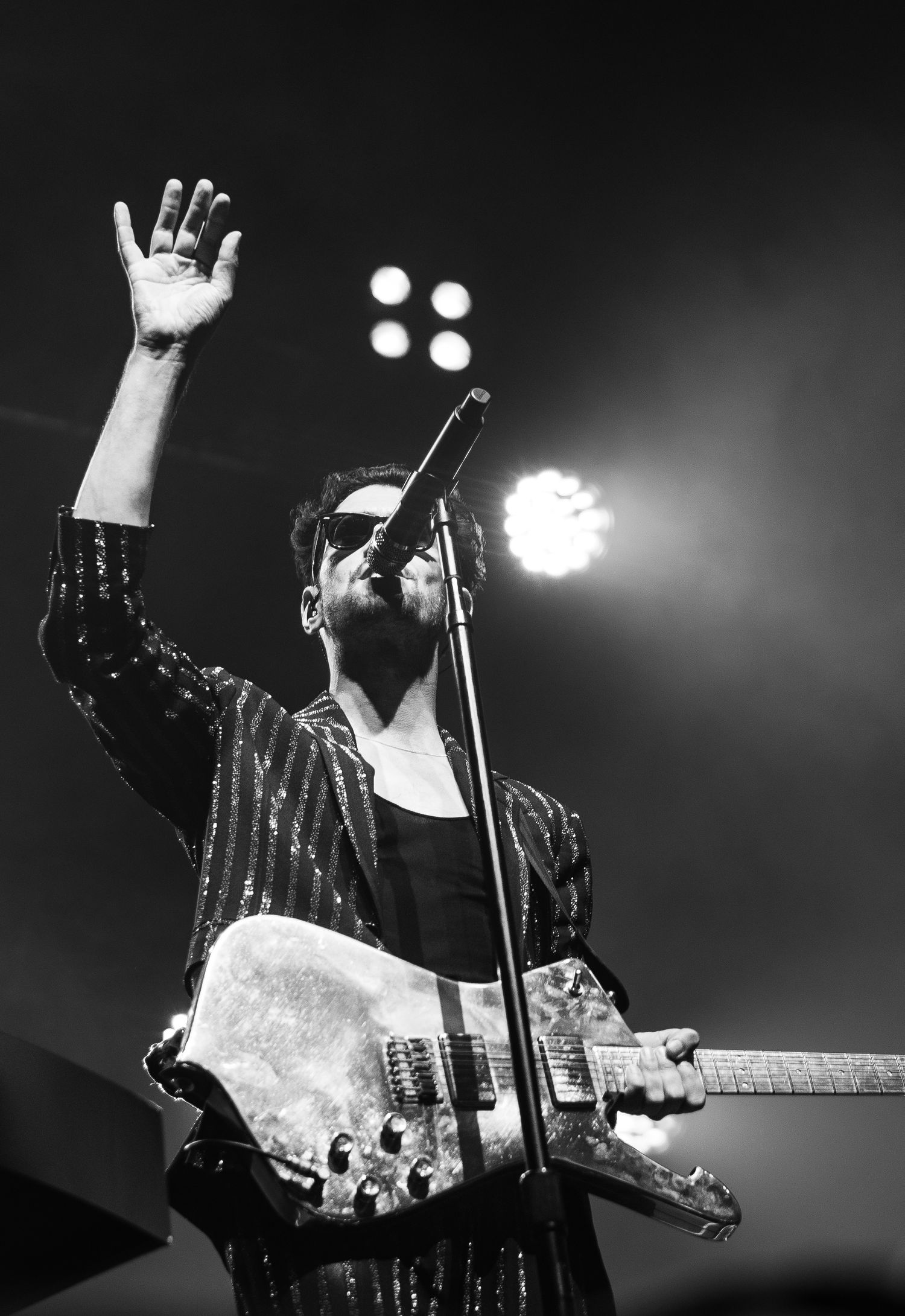Black and white photo of a rock musician playing guitar and raising hand on stage under bright spotlights.