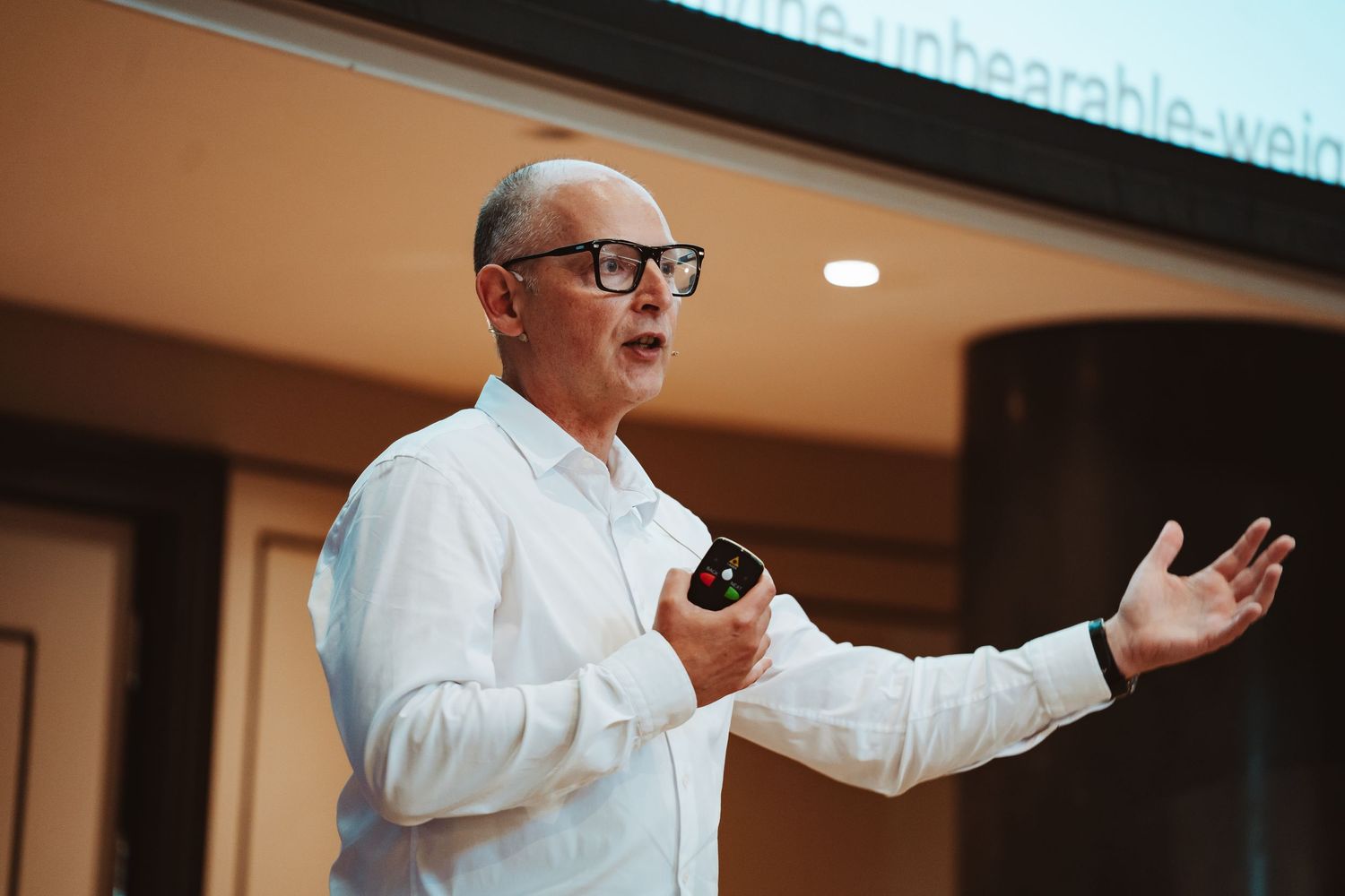 Professional speaker in white shirt and glasses gestures while presenting at indoor conference or event.