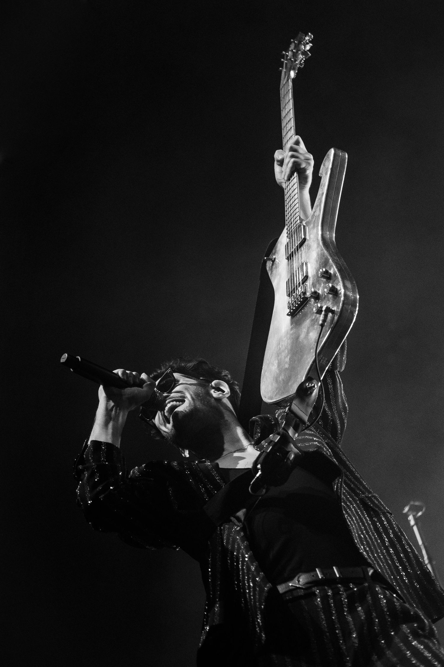 Black and white concert photo of performer holding guitar up while singing into microphone.