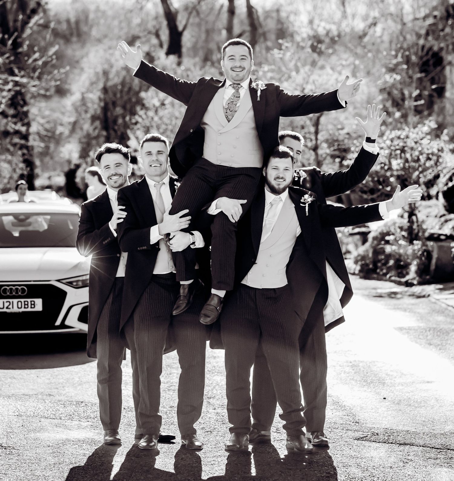 Black and white photo of groom lifted by groomsmen in suits, celebrating outdoors near an Audi car.