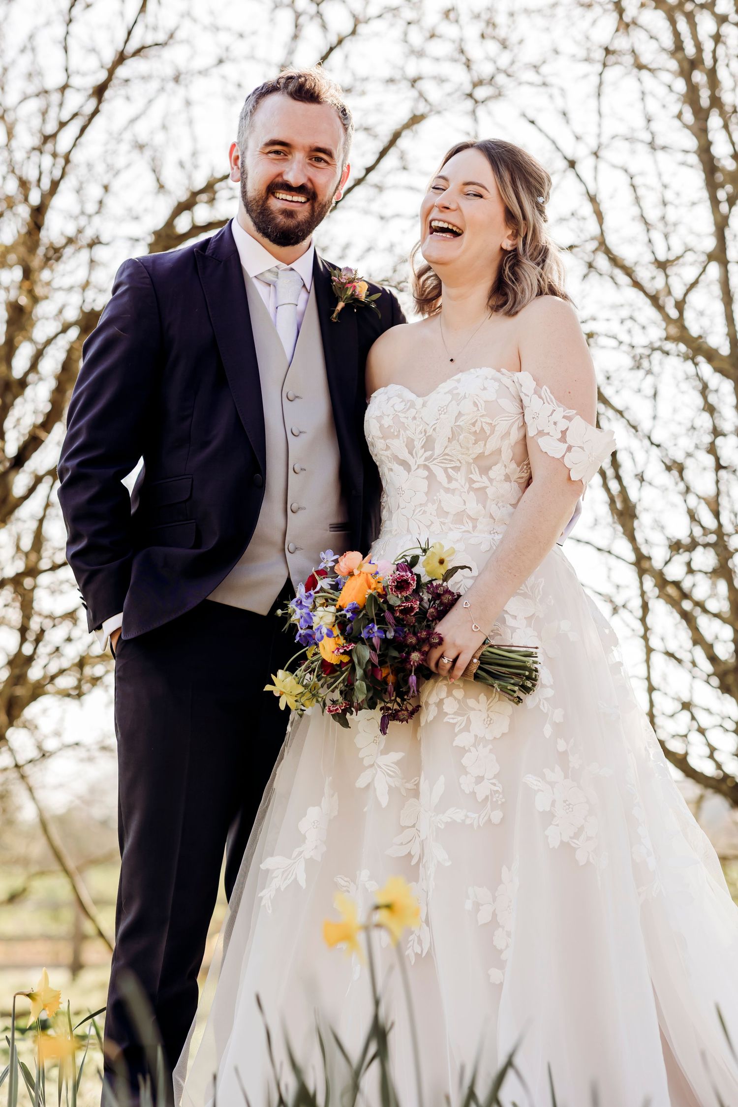 Smiling bride and groom pose outdoors among spring daffodils, bride holding colorful bouquet in lace off-shoulder gown.