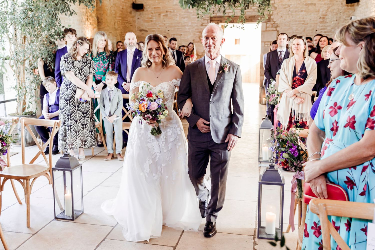 Bride in lace gown walks down the aisle with escort through guests at a rustic barn wedding.