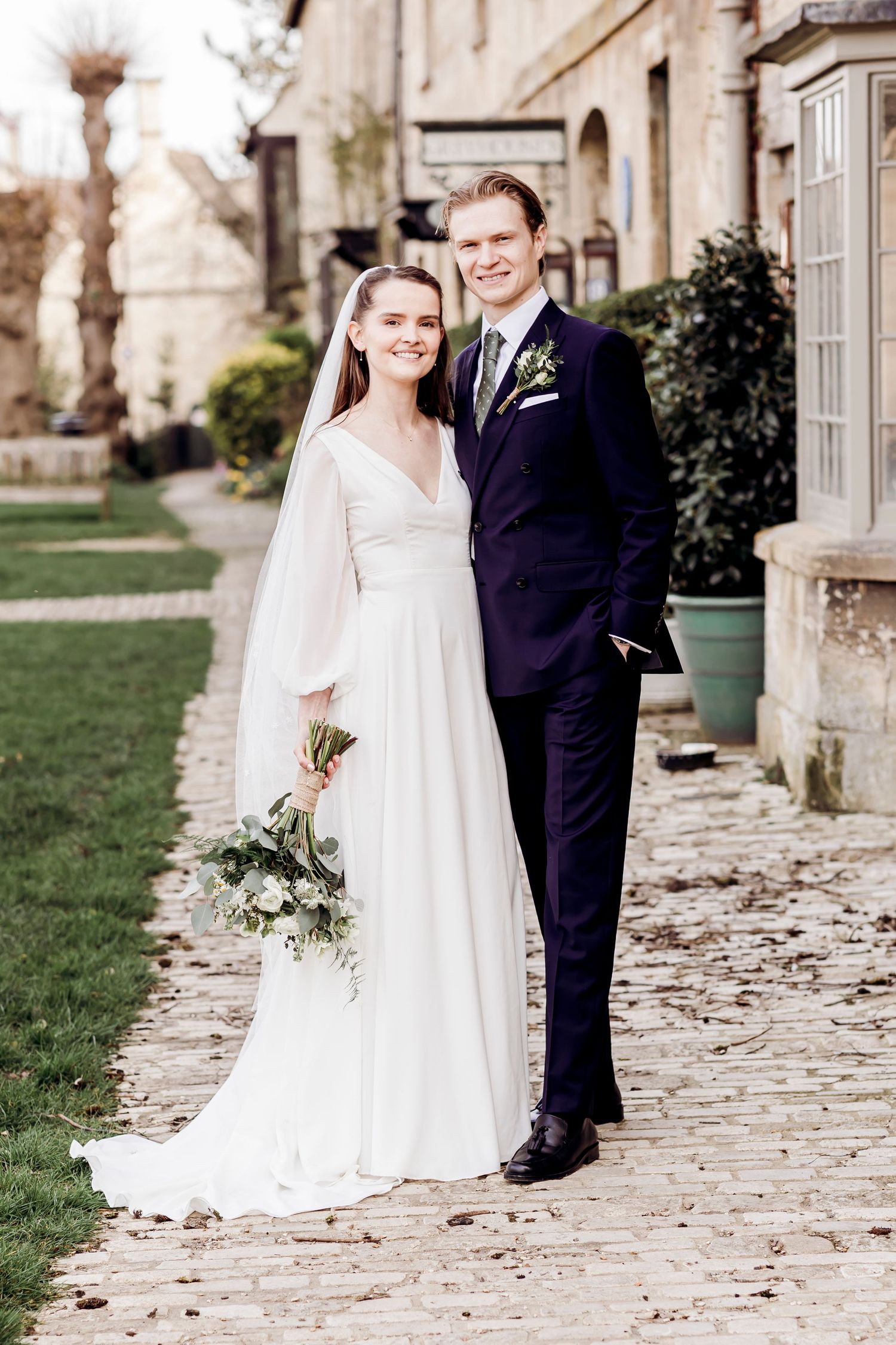 Bride and groom pose together outside historic stone wedding venue holding bouquet.
