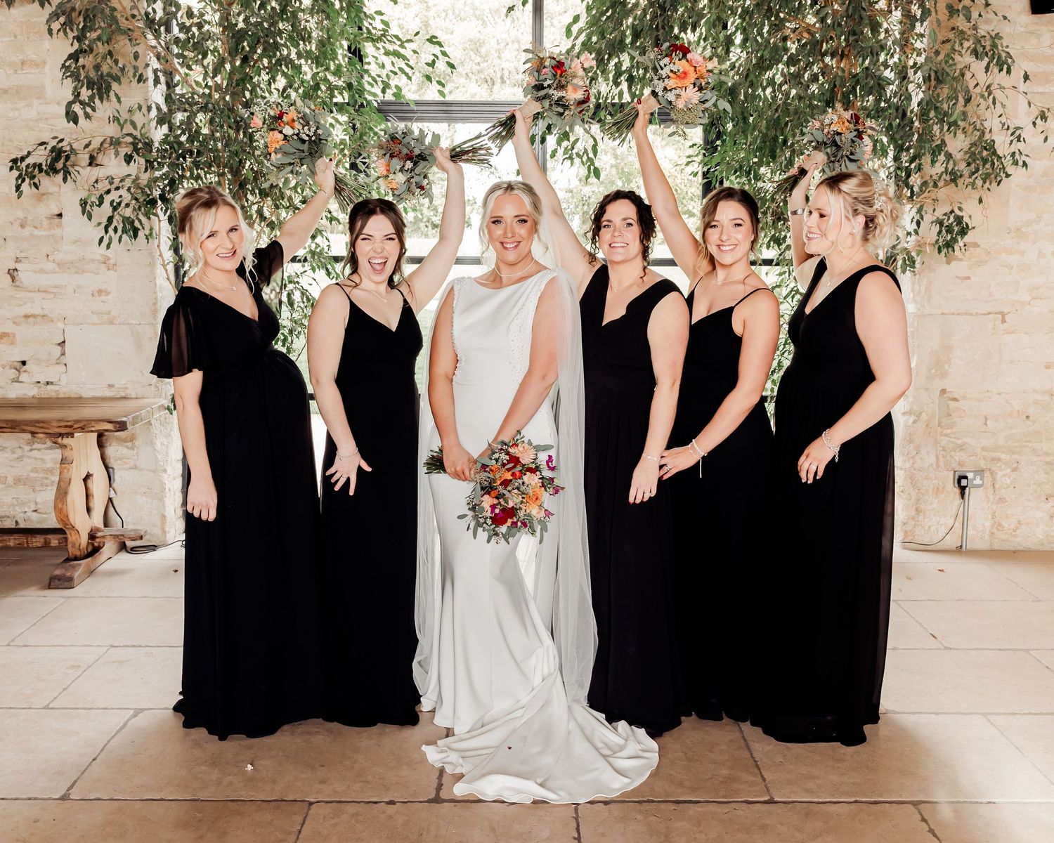 Bride in white gown poses with five bridesmaids in black dresses inside rustic barn with greenery backdrop.