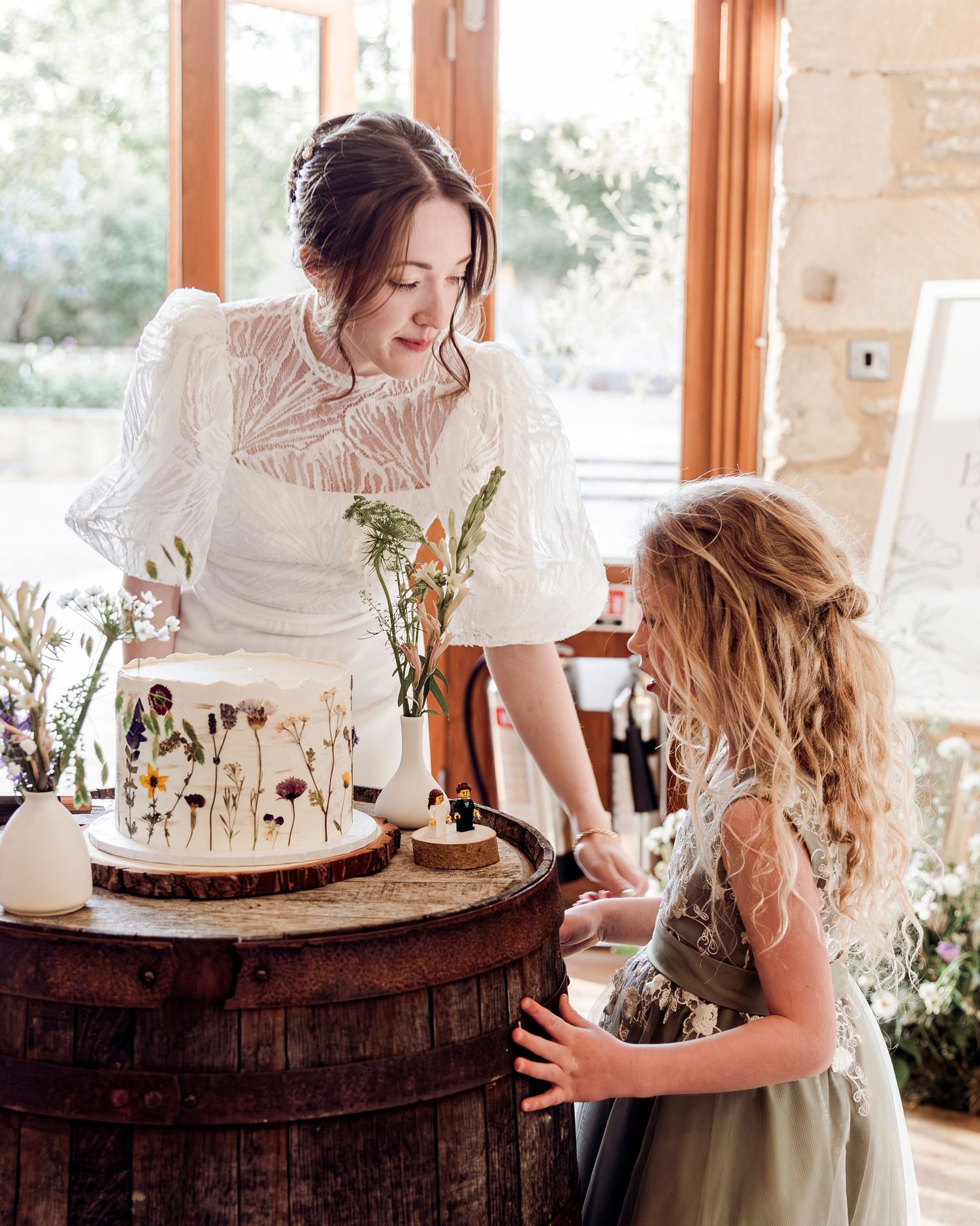 Bride in white lace dress showing decorated floral wedding cake to young flower girl on barrel stand.