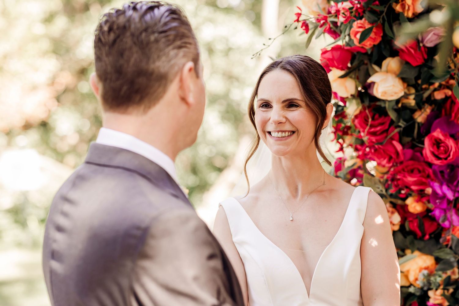 Smiling bride in white V-neck gown exchanges vows before vibrant floral backdrop at outdoor ceremony.